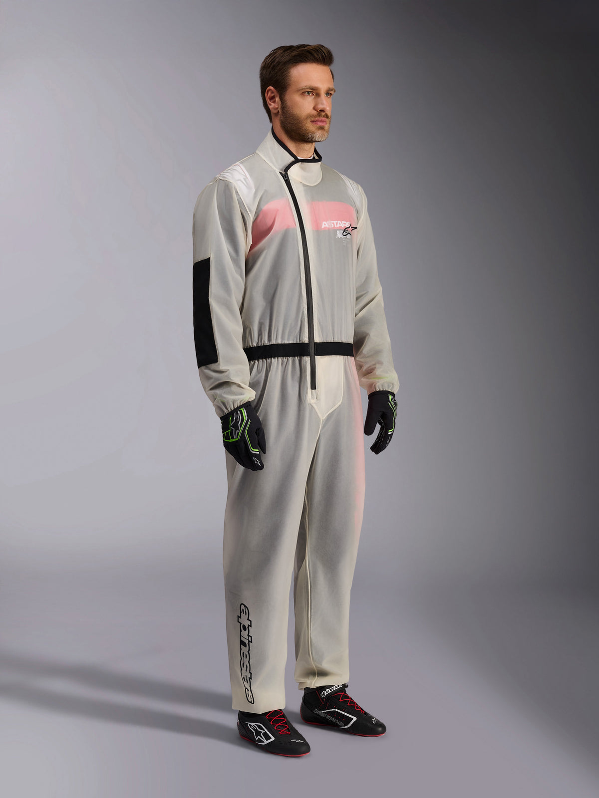 Alpinestars Kart Rain Suit, genomskinlig vattentät regndräkt för karting, halvgenomskinligt material som visar en rosa och vit racingoverall under, med en svart asymmetrisk dragkedja, förstärkt armbågsparti och svart midjeband, bärs med svarta kartinghandskar och racingskor.