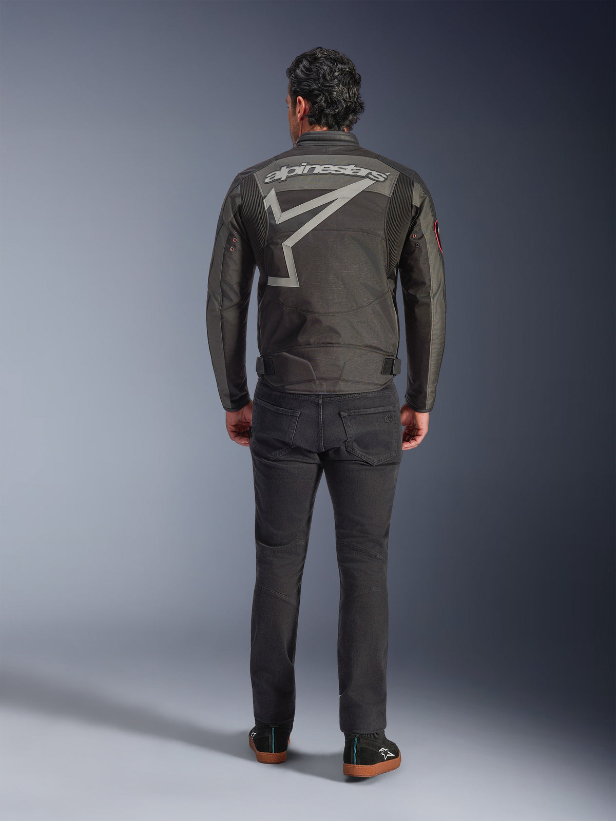 Alpinestars Axton-jacka, motorcykeljacka, svart, antracitgrå, röd och orange, vy bakifrån som visar en stor grå Alpinestars-logotyp och stjärngrafik, dragspelspaneler på axlarna, bärs med svarta byxor och vardagliga svarta skor.