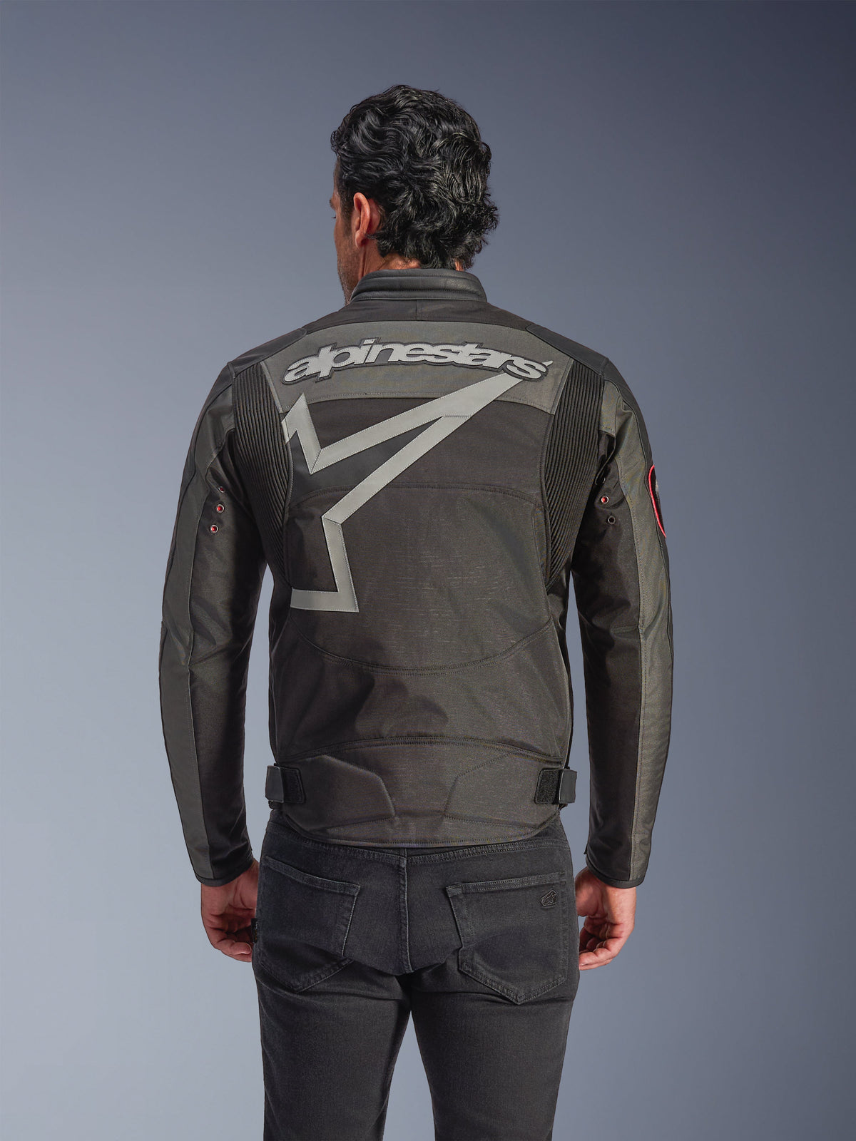 Alpinestars Axton jacka, motorcykeljacka för herrar, Black Anthracite Red Tangerine, vy bakifrån med ett stort grått Alpinestars-ordmärke och stiliserad stjärngrafik, dragspelspaneler på axlarna och vintageinspirerade metallöljetter på ärmarna.