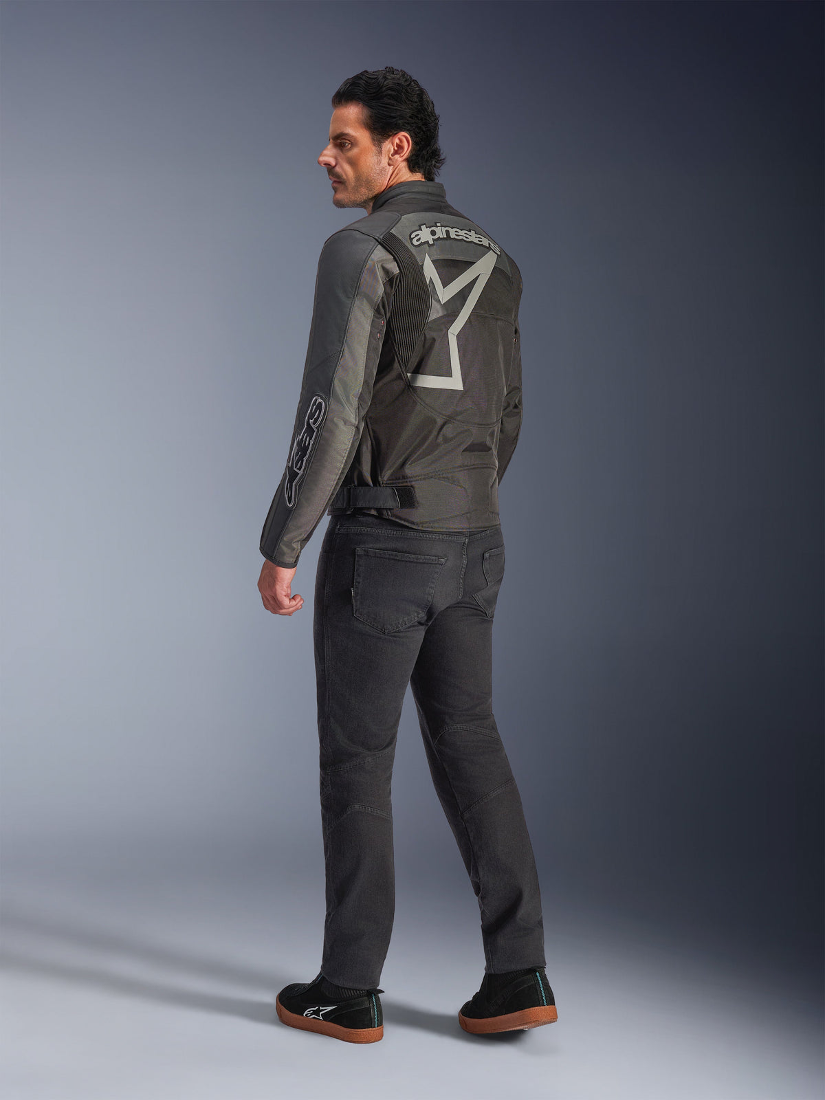 Alpinestars Axton-jacka, motorcykeljacka för herrar, svart och antracitgrå, vy bakifrån med en stor grå omslutande logotyp och dragspelspaneler på axlarna, bärs med mörka mc-byxor och svarta sneakers, urban modern stil med racingarv.