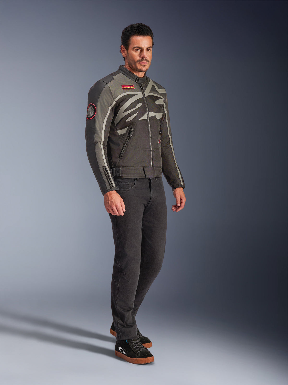 Manlig modell bär Alpinestars Axton Jacket i Black Anthracite Red Tangerine, en vintageinspirerad motorcykeljacka med en omslutande logotyp och röda detaljer, matchad med svarta byxor och svarta MC-sneakers, urban samtida motorcykelutrustning.