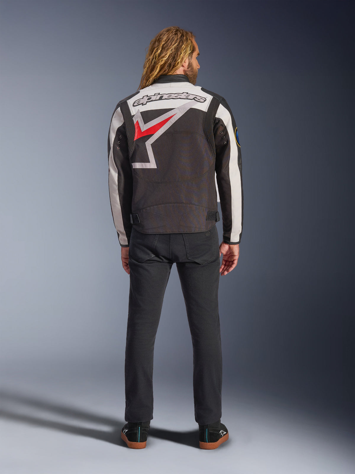 Alpinestars Axton Jacket, Motorcycle Jacket, Black Off White and Bright Red, back view on a model, featuring a large Alpinestars star logo on the back, off-white sleeve panels, worn with black pants, vintage-inspired racing teamwear design