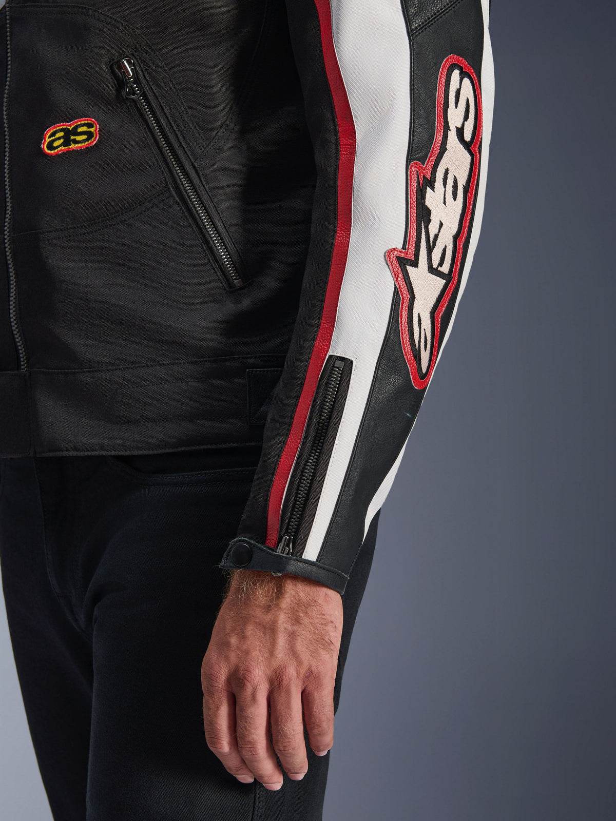 Alpinestars Axton Jacket, close-up of sleeve and torso, Black with Off White and Bright Red accents, featuring large embroidered Alpinestars logo on the arm, vertical racing stripes, zippered cuff with snap, and rugged textile construction.