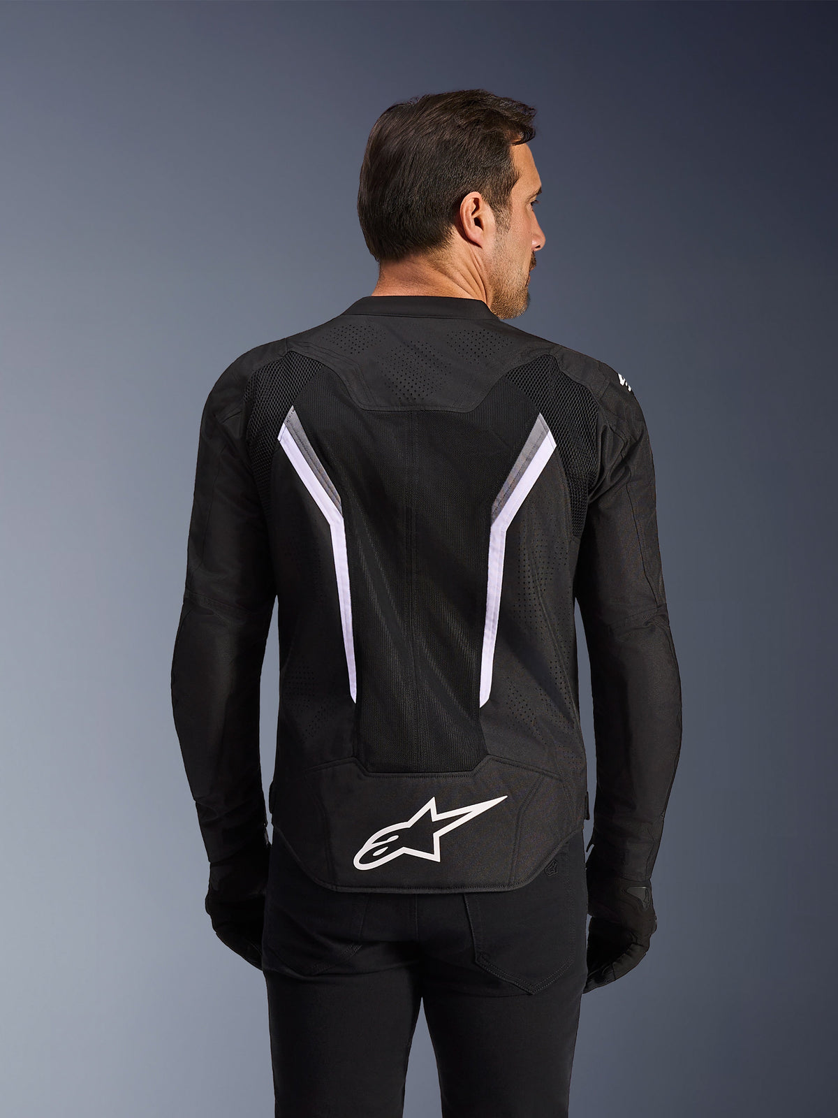 Alpinestars AST V3 Air Jacket, Motorcycle Jacket, Black with White and Dark Gray accents, back view showing extensive mesh ventilation panels, large white V-shaped stripes, and white Alpinestars logo on the lower back, worn with black gloves and pants