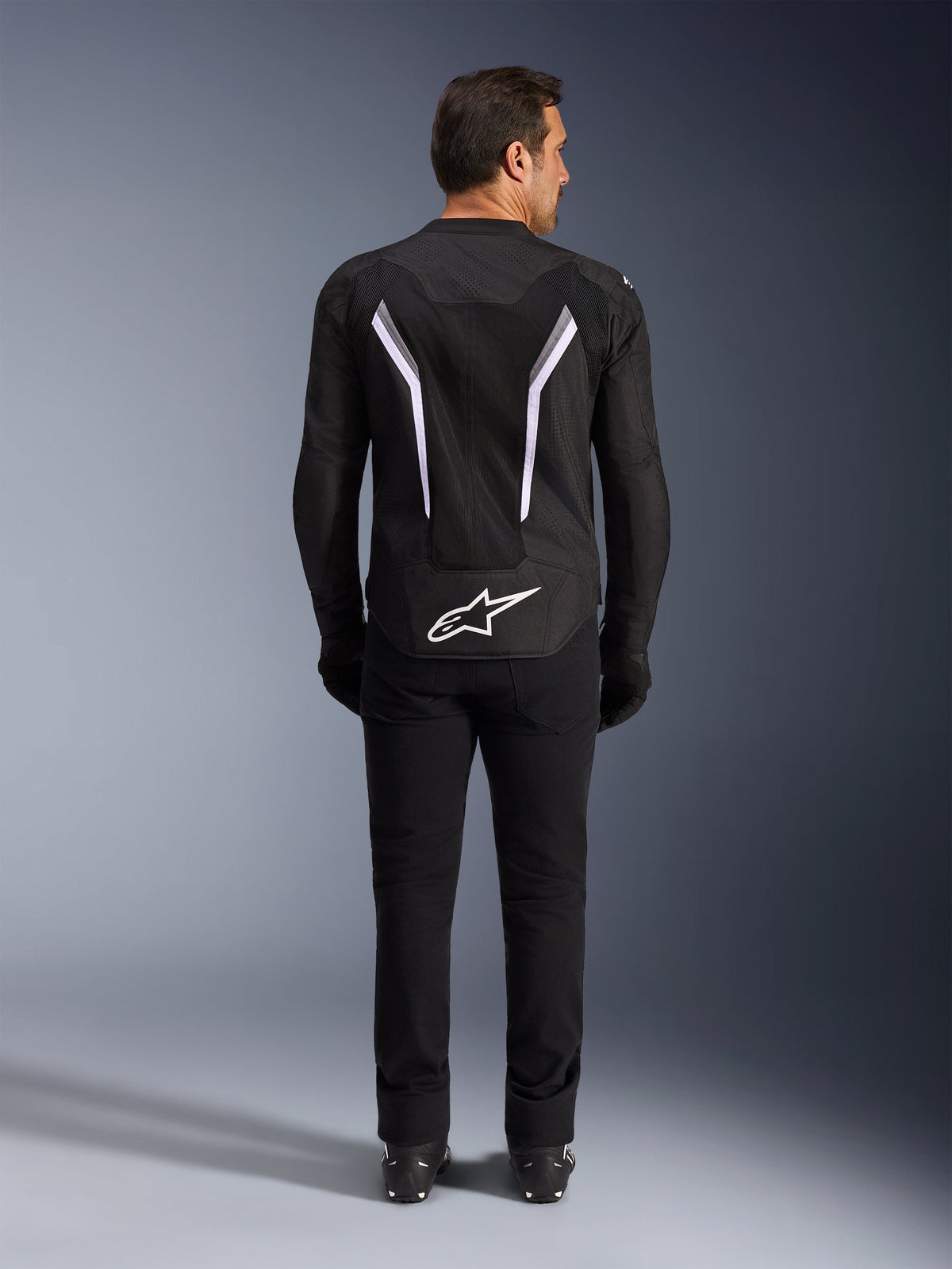 Alpinestars AST V3 Air Jacket, Back View, Black and Dark Gray with White accents, featuring large white Astar logo on the tail, reflective stripes on the back, and mesh textile construction, worn with black pants and boots for a sport riding look