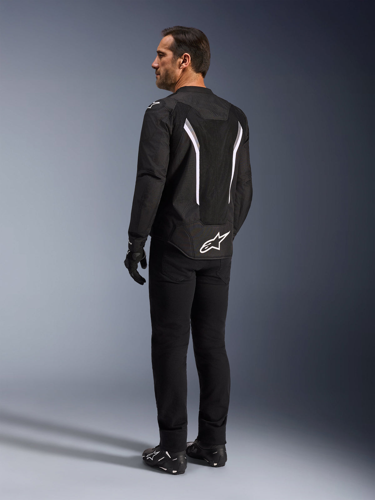 Alpinestars AST V3 Air Jacket, Black and White motorcycle jacket, back view on model, featuring mesh inserts for airflow and white logo on the lower back, worn with black technical riding gear