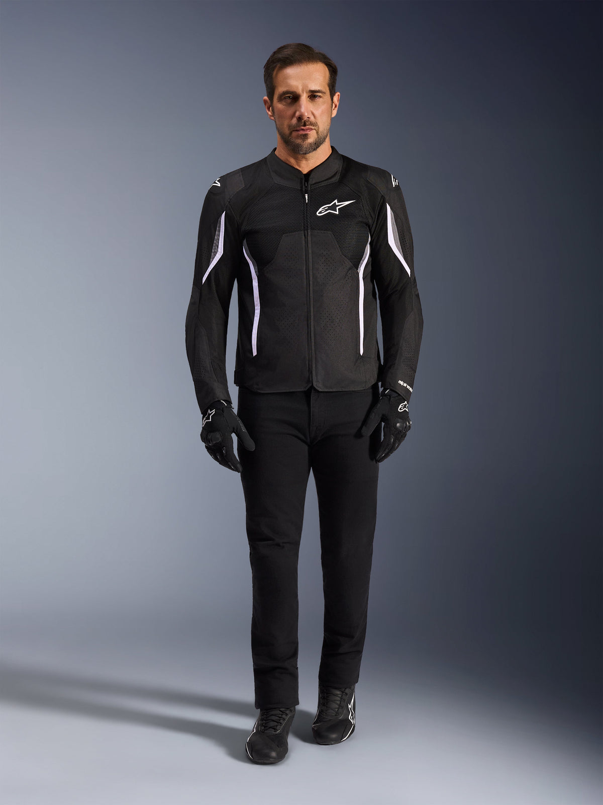 Male model wearing the Alpinestars AST V3 Air Jacket in Black with white accents, featuring mesh ventilation panels and a sporty fit, paired with black technical riding pants, gloves, and boots, front view, technical motorcycle gear for warm-weather riding.