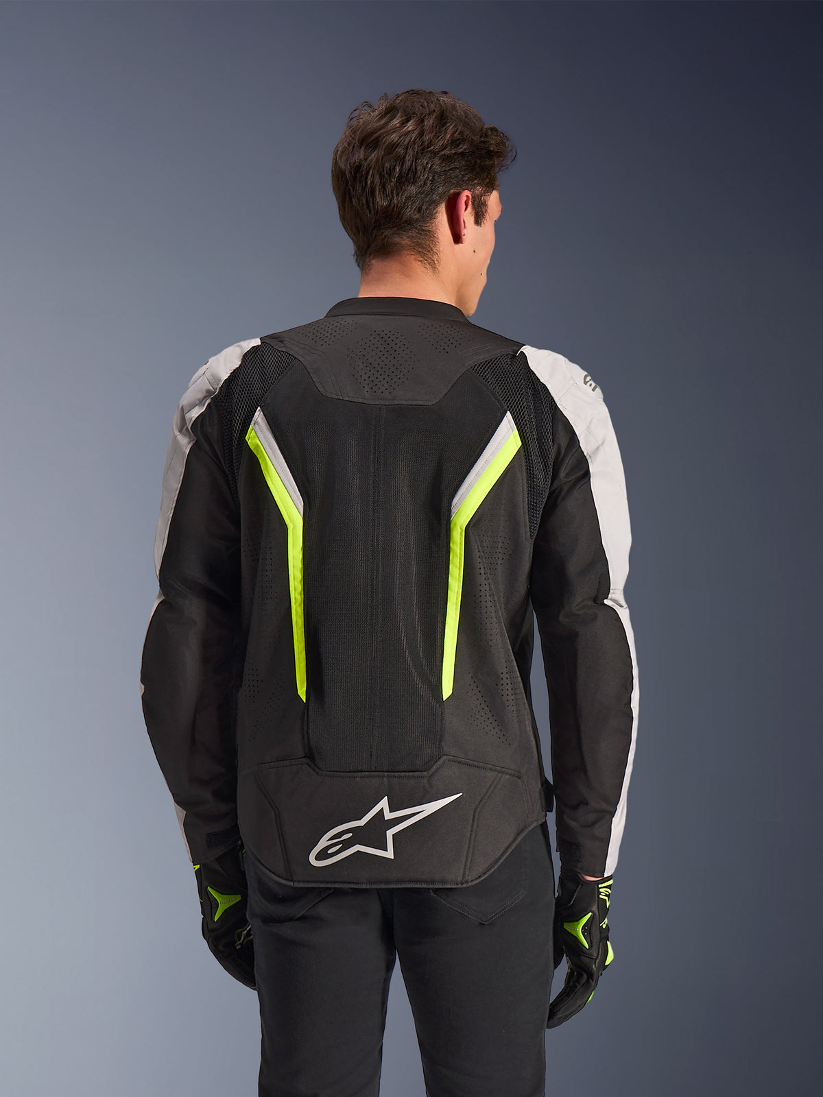 Alpinestars AST V3 Air Jacket, Svart Silver Gul Fluo, vy bakifrån på modell, mc-jacka i textil med dubbla ventilationspaneler i tech mesh, vertikala neongula ränder, silverfärgade ärmar och en vit Alpinestars-logotyp på nedre delen av ryggen.