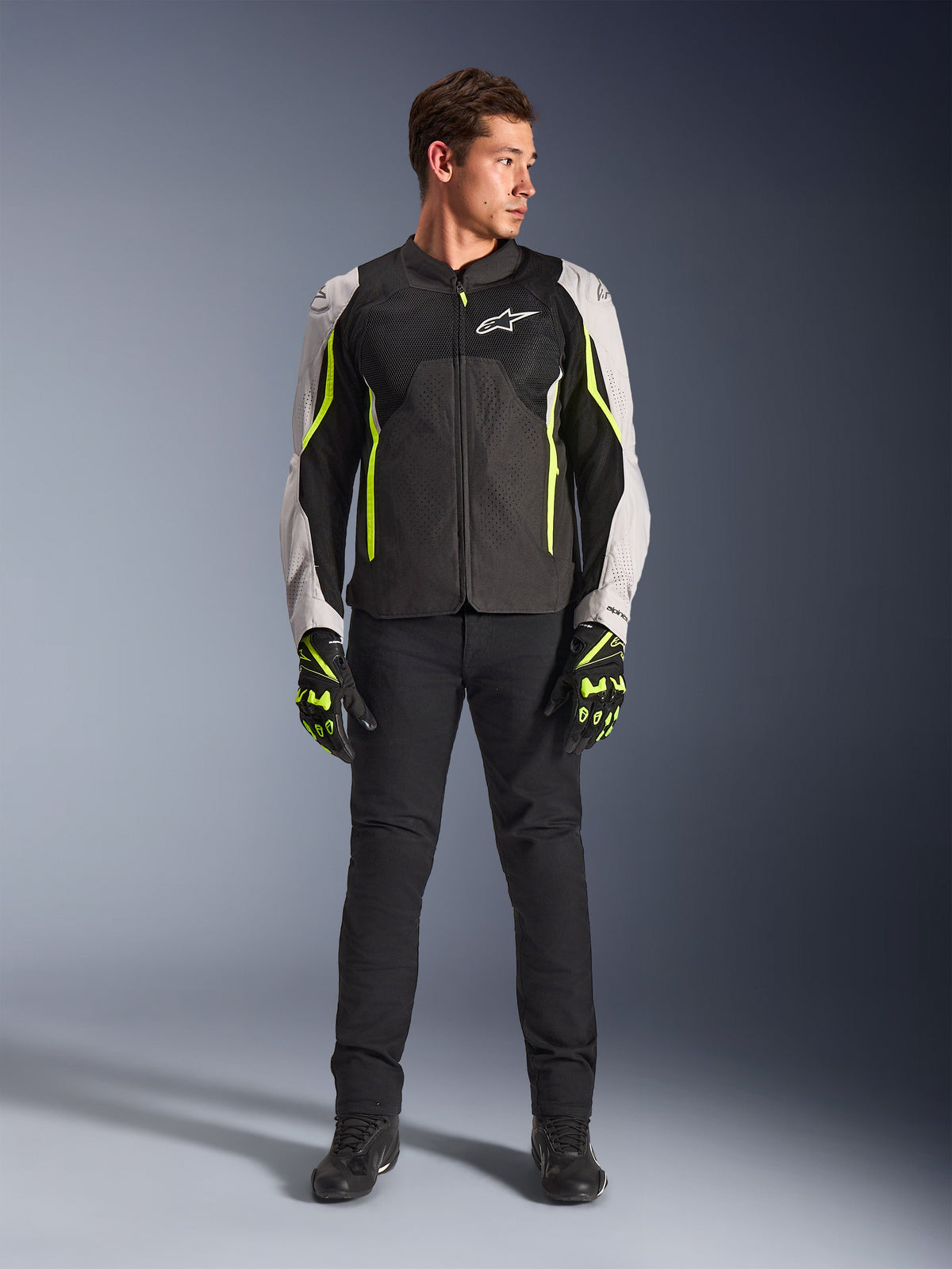 Manlig modell som bär Alpinestars AST V3 Air-jacka i Black Silver Yellow Fluo, med ventilationspaneler i mesh och fluorescerande detaljer, kombinerat med svarta mc-byxor och matchande motorcykelhandskar, sportig prestandautrustning.