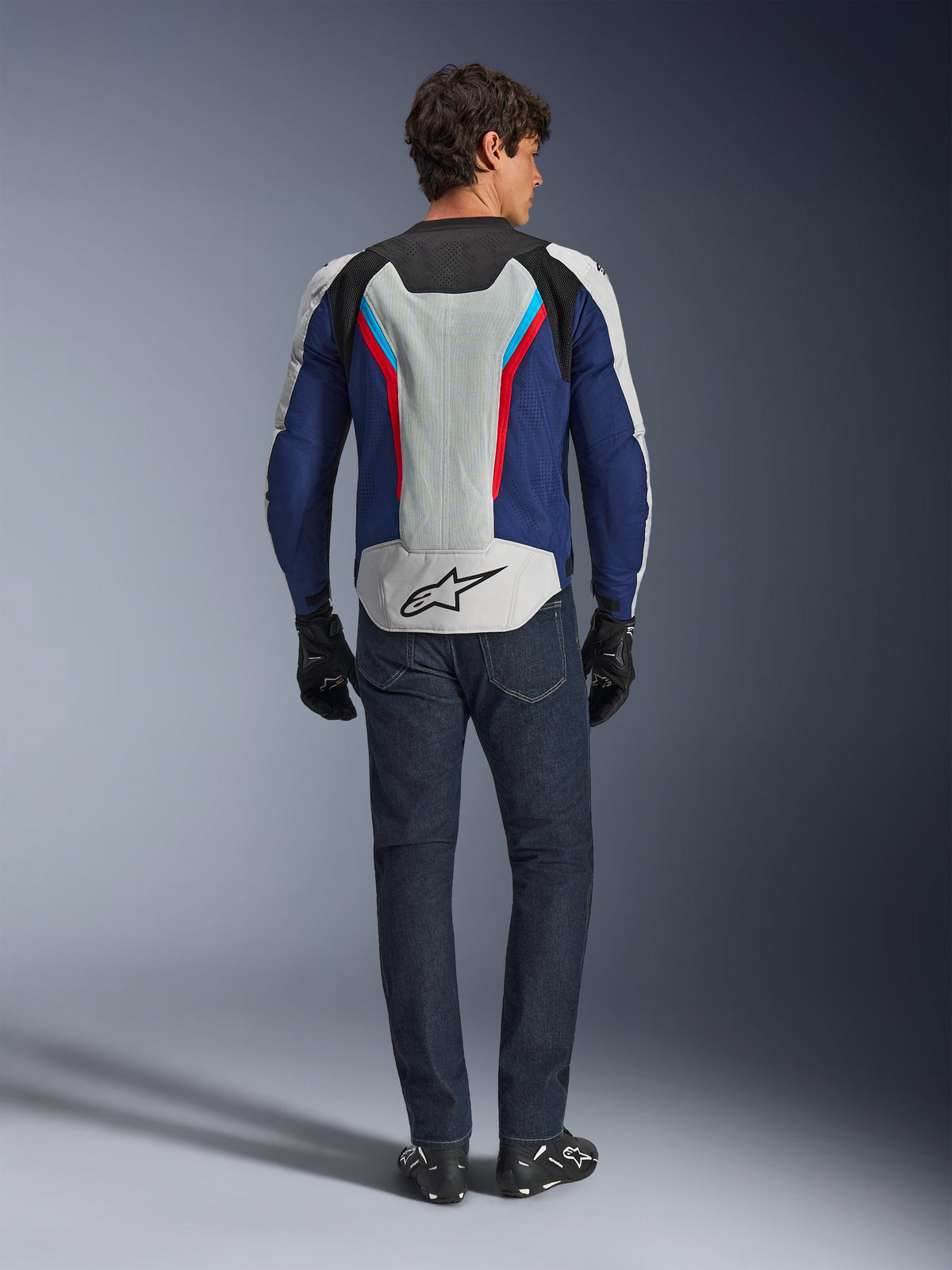 Alpinestars AST V3 Air Jacket, sedd bakifrån, Silver Deep Blue och Bright Red, med en stor ventilationspanel i mesh på ryggen med Alpinestars-logotypen, bärs med mörkblå jeans och svarta handskar, motorcykelutrustning i sportpassform för varmt väder