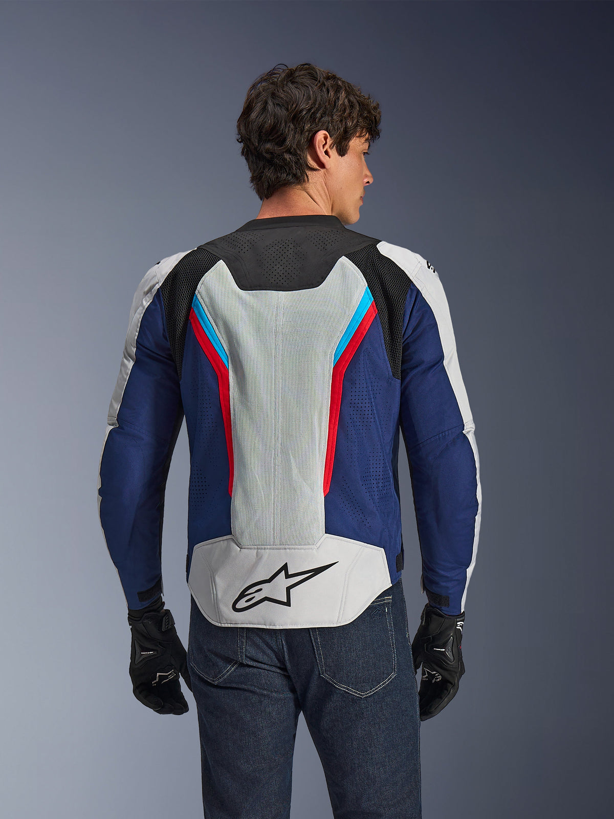 Alpinestars AST V3 Air Jacka, motorcykeljacka, silver, mörkblå och klarröd, sedd bakifrån buren av en förare, med tekniska meshpaneler och laserperforeringar för ventilation, svart stjärnlogotyp på nedre delen av ryggen, sportoptimerad design för varmt väder