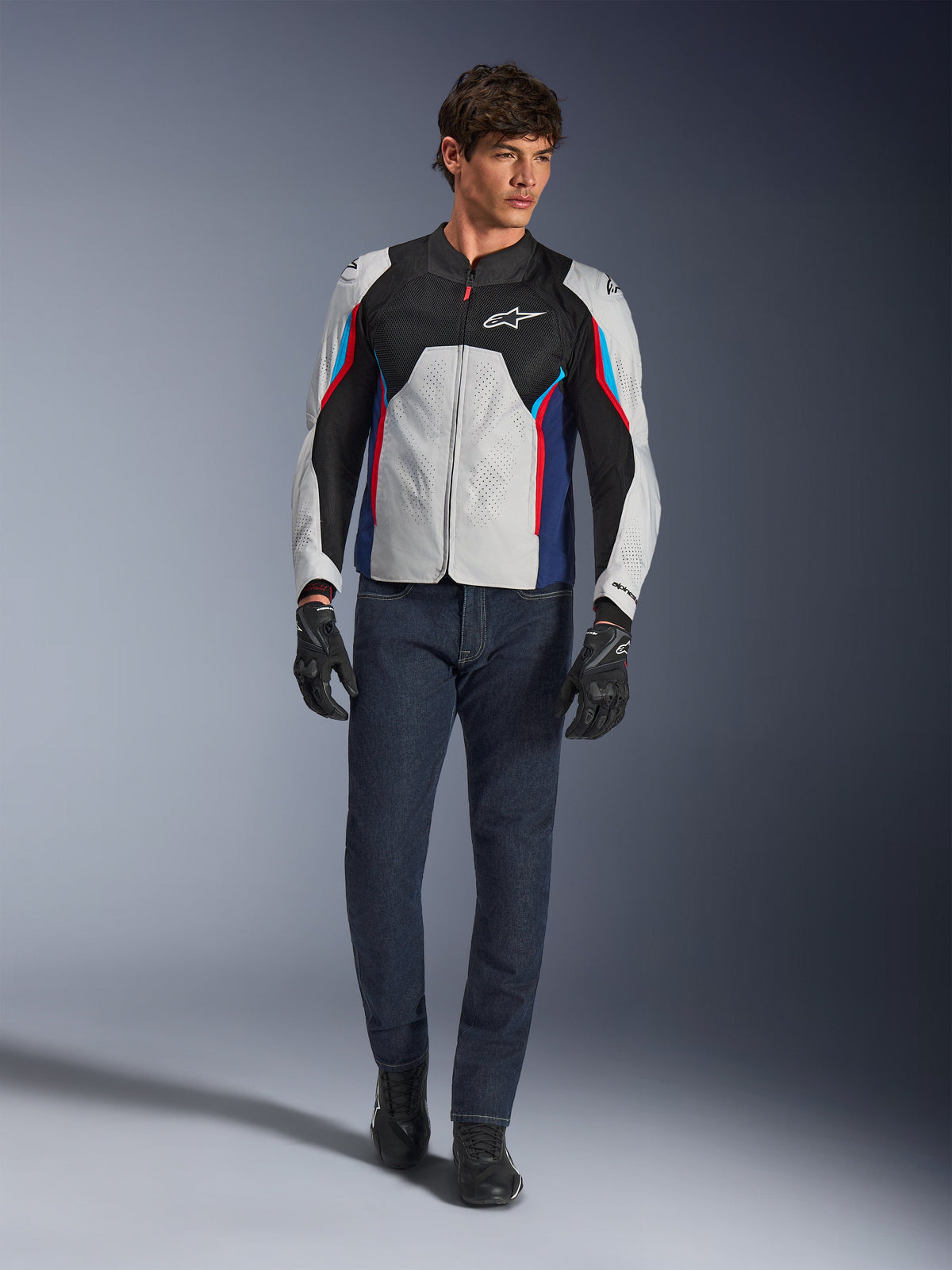 Manlig modell som bär Alpinestars AST V3 Air Jacket i Silver Deep Blue Bright Red, med meshventilation och perforerade paneler, matchat med mörka mc-jeans i denim och svarta handskar, sportoptimerad motorcykelutrustning för varmt väder.
