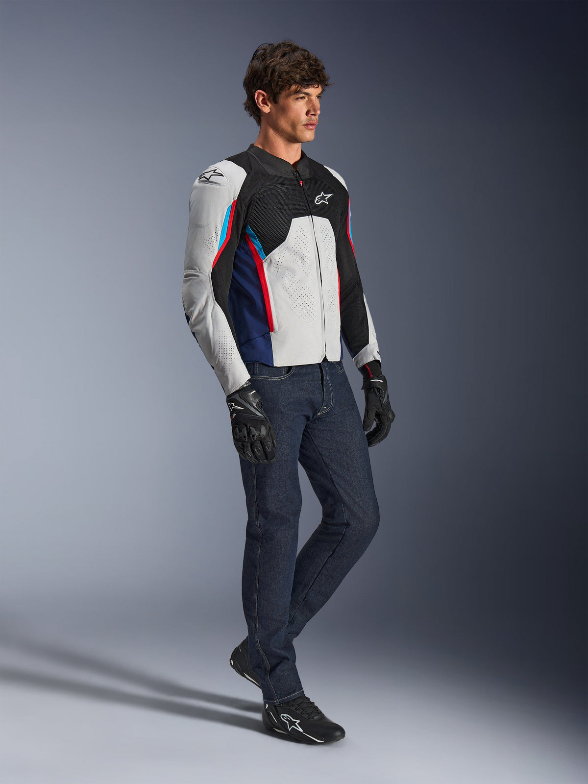 Alpinestars AST V3 Air-jacka, motorcykeljacka, silver, mörkblå och klarröd, bärs med mörka denimjeans och svarta körhandskar, med ventilerade meshpaneler och en sport-fit-design för körning i varmt väder