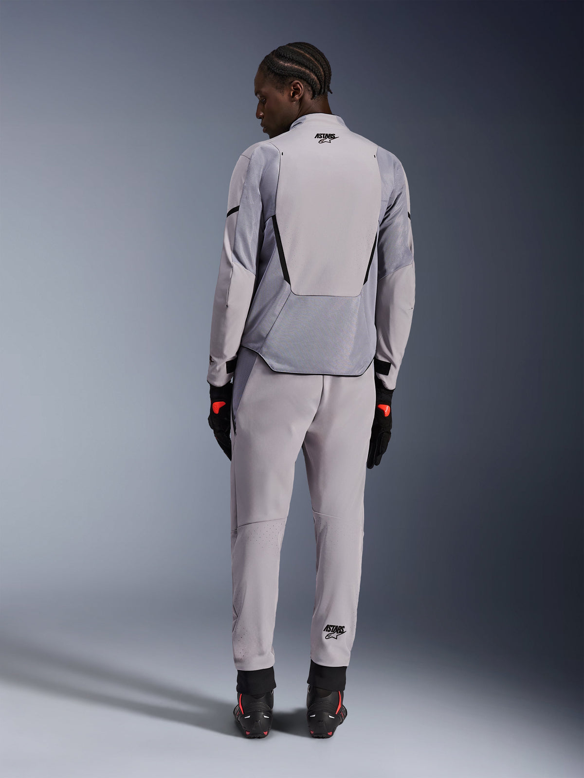 Alpinestars Aeroshell Airflow-jacka, Mid Gray Anthracite, sedd bakifrån på en modell, med tekniska meshpaneler och laserperforerade ventilationszoner, buren med matchande gråa byxor och svart motorcykelutrustning, sportskuren design för hög prestanda.