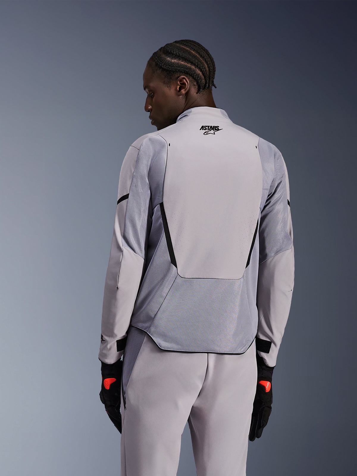 Alpinestars Aeroshell Airflow-jacka i Mid Gray Anthracite, ryggvy som visar ventilerande meshpaneler och laserperforeringar, teknisk stretch-softshellkonstruktion med en förlängd bakpanel, bärs med matchande gråa byxor och svarta handskar för en sportig look.