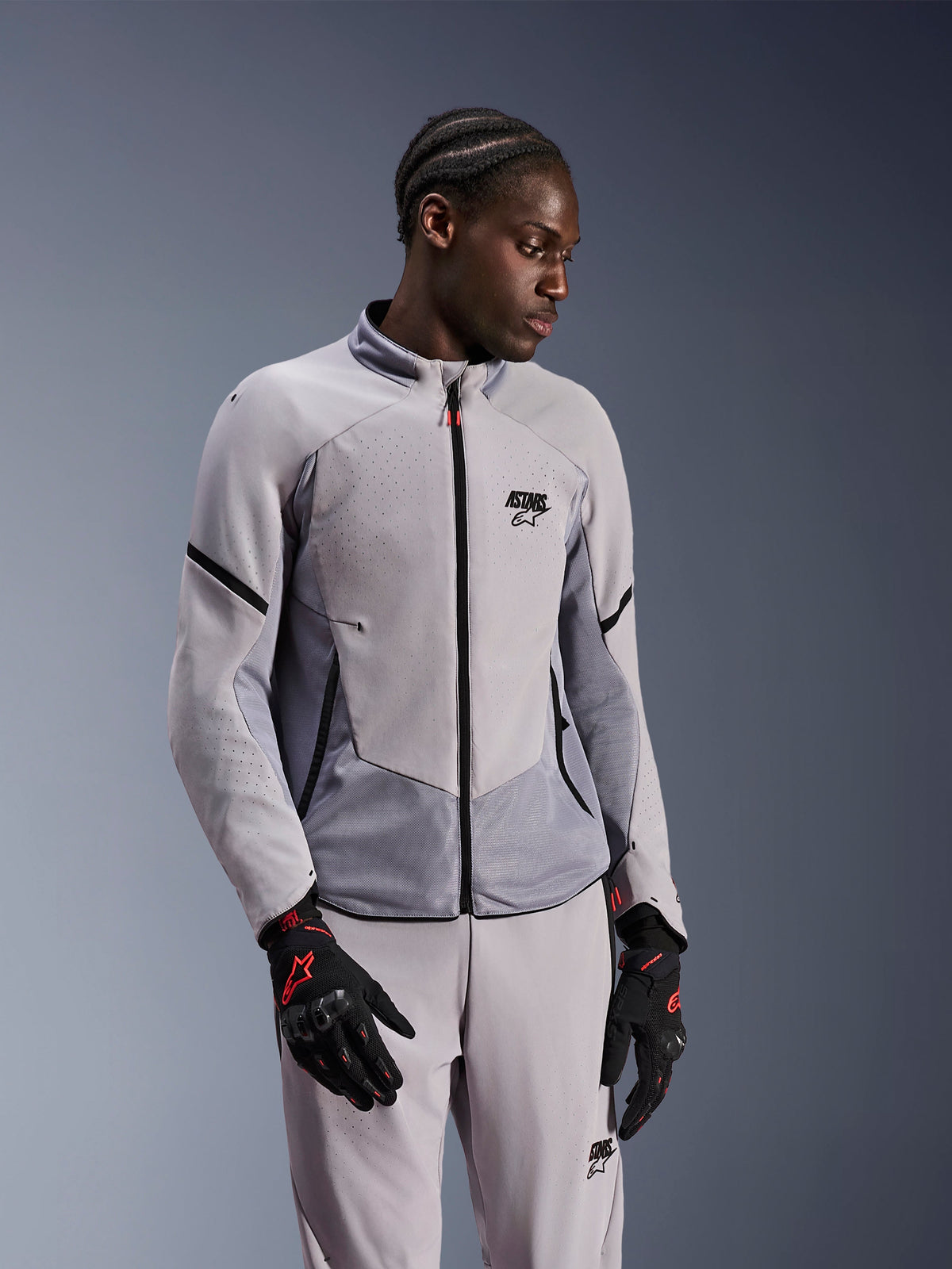 Alpinestars Aeroshell Airflow-jacka, Mid Gray Anthracite, bärs av en modell med matchande textilbyxor och svarta handskar med röda detaljer, med laserperforerade zoner och meshpaneler för ventilation, strömlinjeformad sport-fit-design för motorcykelkörning