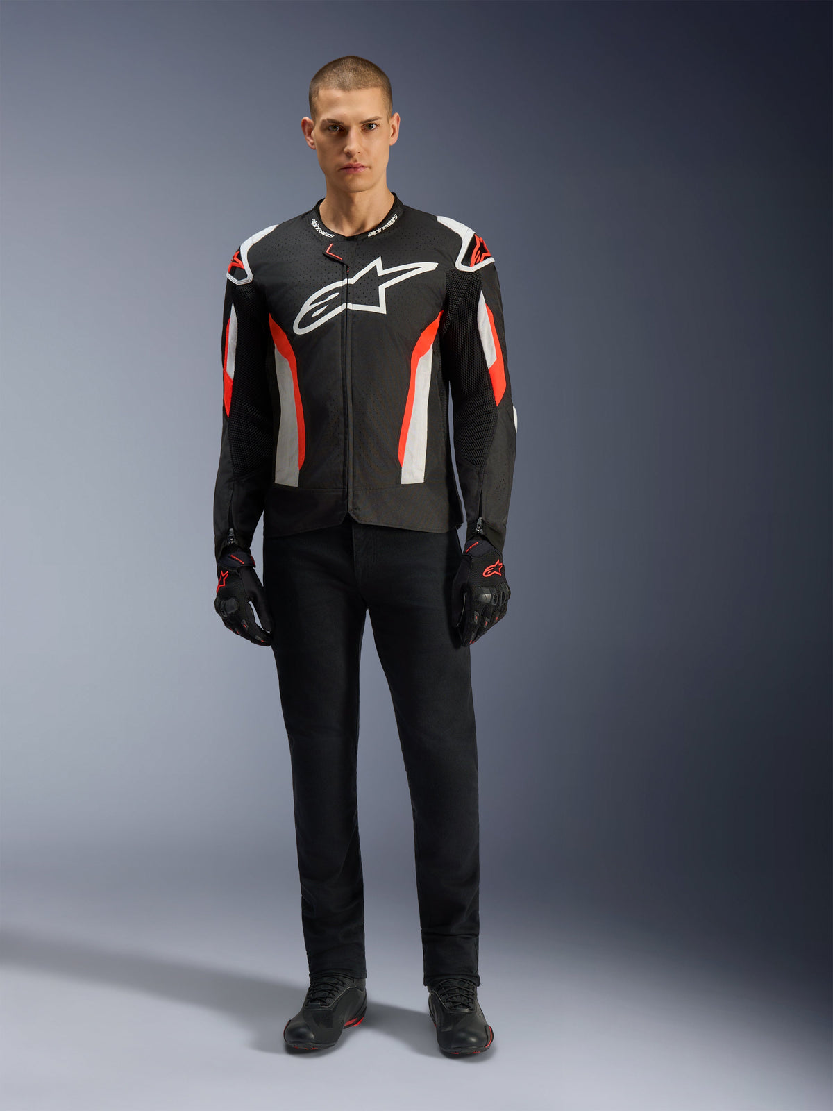 Male model wearing the Alpinestars T-GP Air Jacket in Black Red Fluo, textile motorcycle jacket with white and bright red accents, large white Alpinestars logo on the chest, featuring mesh ventilation panels, paired with black technical pants and riding gloves, sport-cut gear