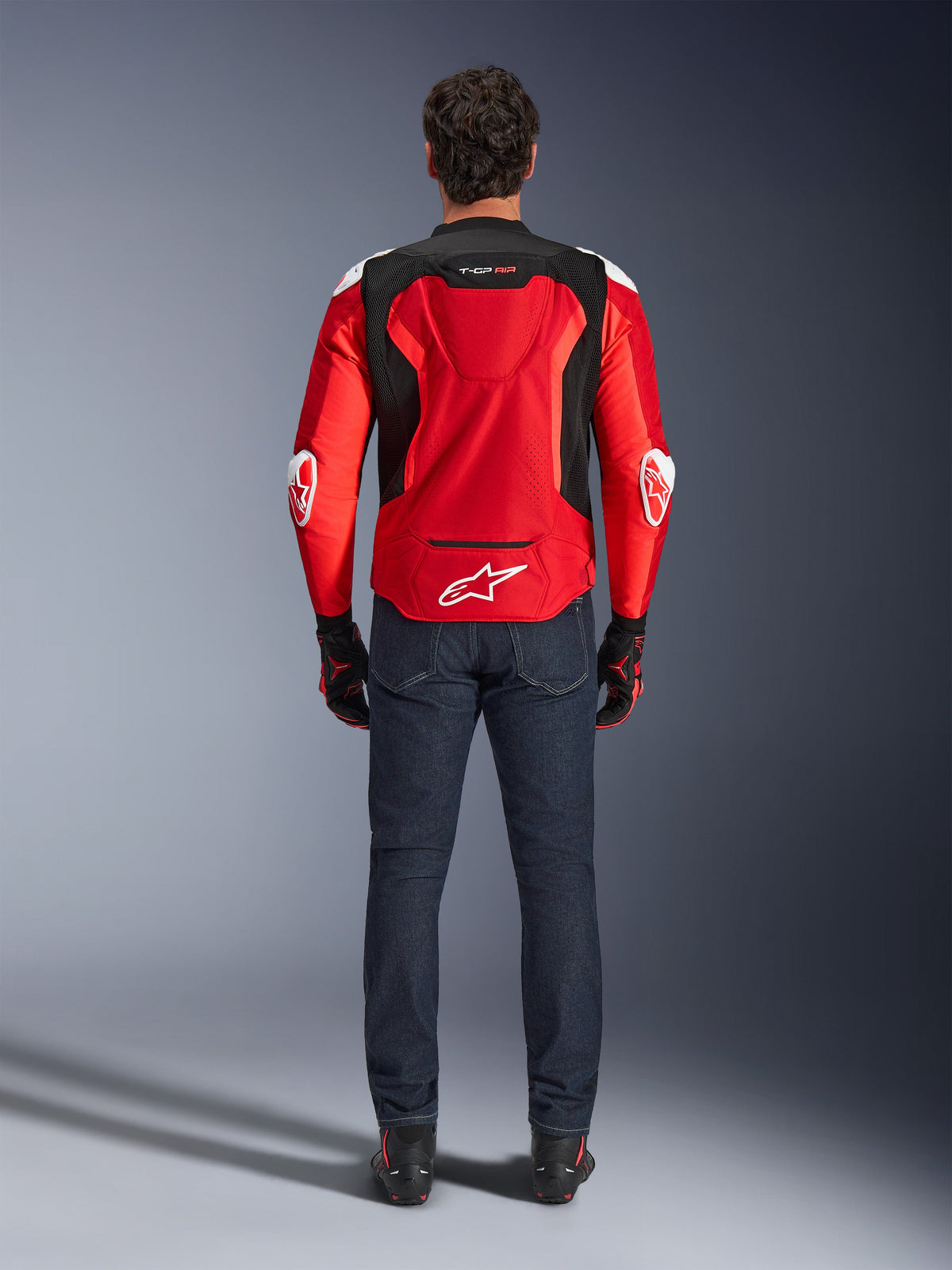 Alpinestars T-GP Air-jacka, vy bakifrån, klaröd och svart, med vit logotyp på nedre delen av ryggen och 'T-GP AIR'-märkning, buren med mörkblå denimjeans och svarta och röda körhandskar, motorcykelutrustning för sportprestanda