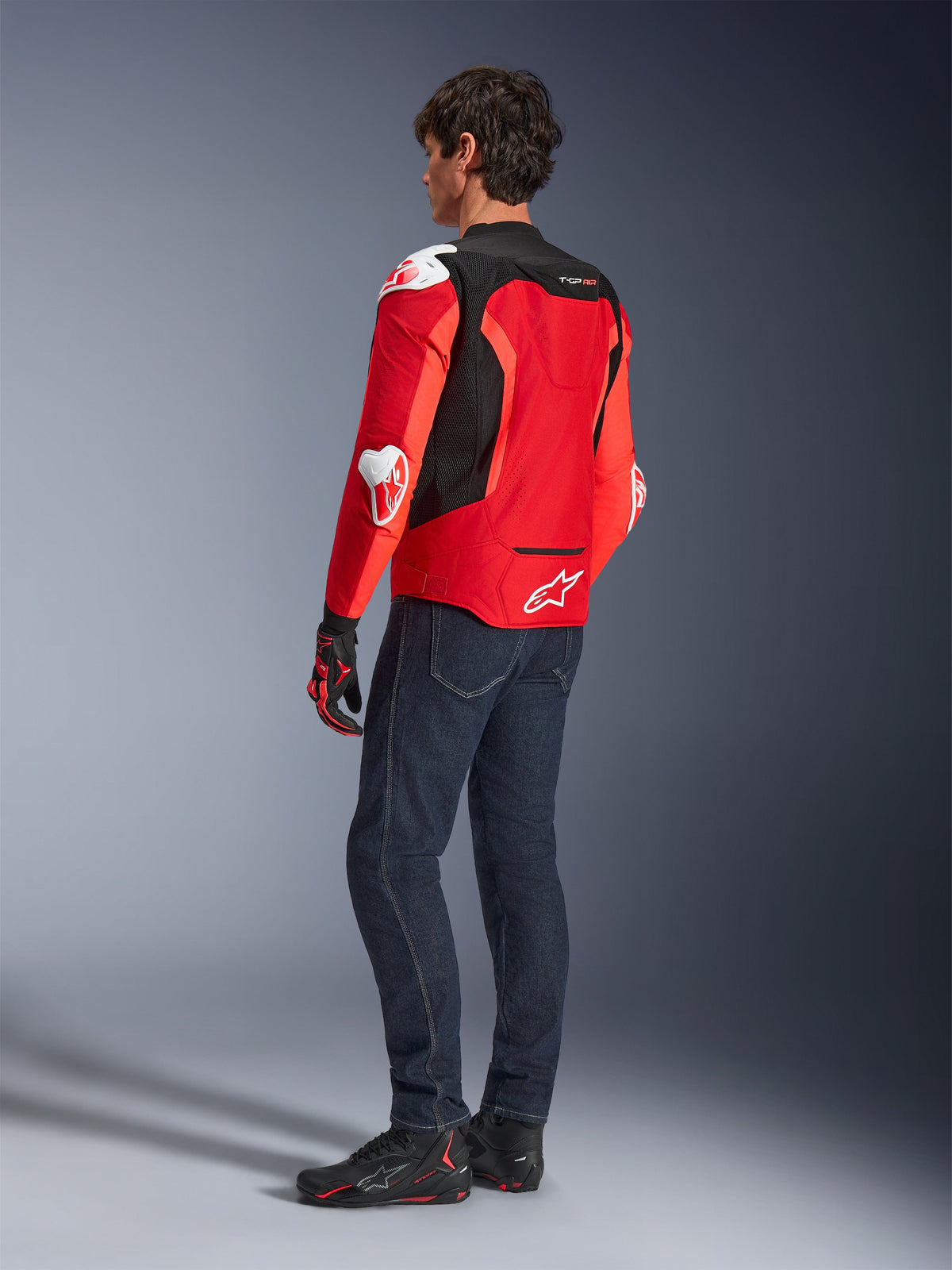 Alpinestars T-GP Air-jacka, ryggvy av en man klädd i en klar röd och neonröd textiljacka med svarta meshpaneler och vita sliders, kombinerat med mörkblå jeans och svarta mc-skor, sportig motorcykelutrustning