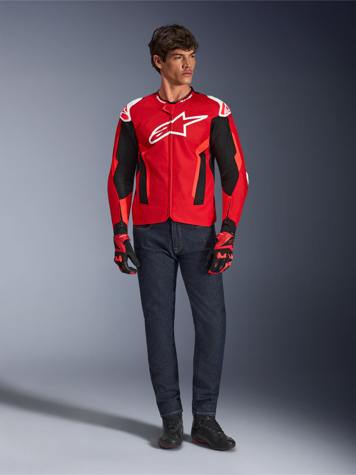Alpinestars T-GP Air-jacka, Bright Red Red Fluo Black, manlig modell i fullständig motorcykeloutfit med mörkblå jeans och matchande handskar, sportig textiljacka med perforerade meshpaneler och vit Astars-logotyp, studiobild.