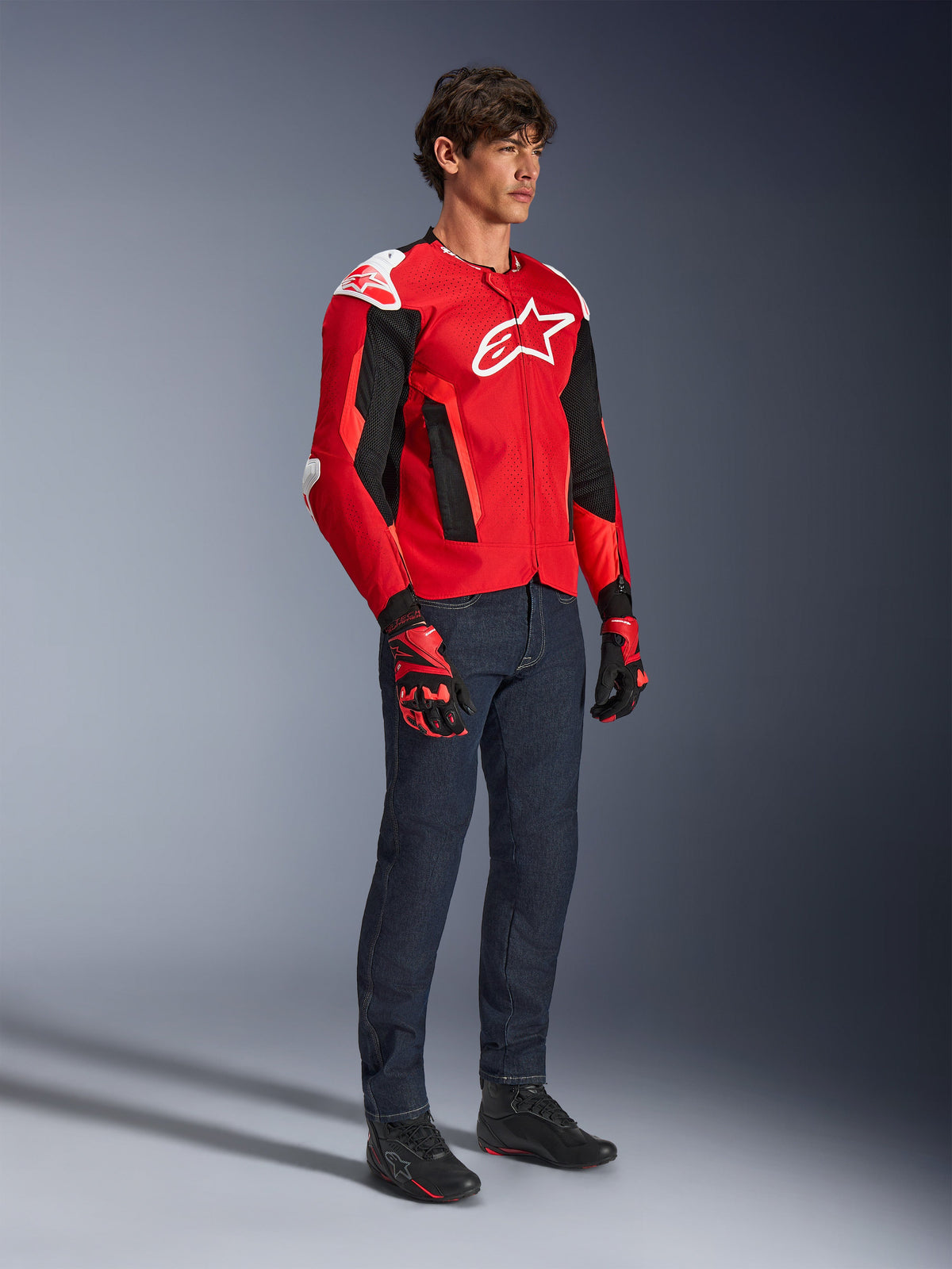 Alpinestars T-GP Air Jacket, sportmotorcykeljacka, klart röd och svart med vita detaljer, bärs av en modell med mörkblå jeans och röda racinghandskar, med meshpaneler för högt luftflöde och vita axelsliders, aerodynamisk sportpassform för körning i varmt väder.