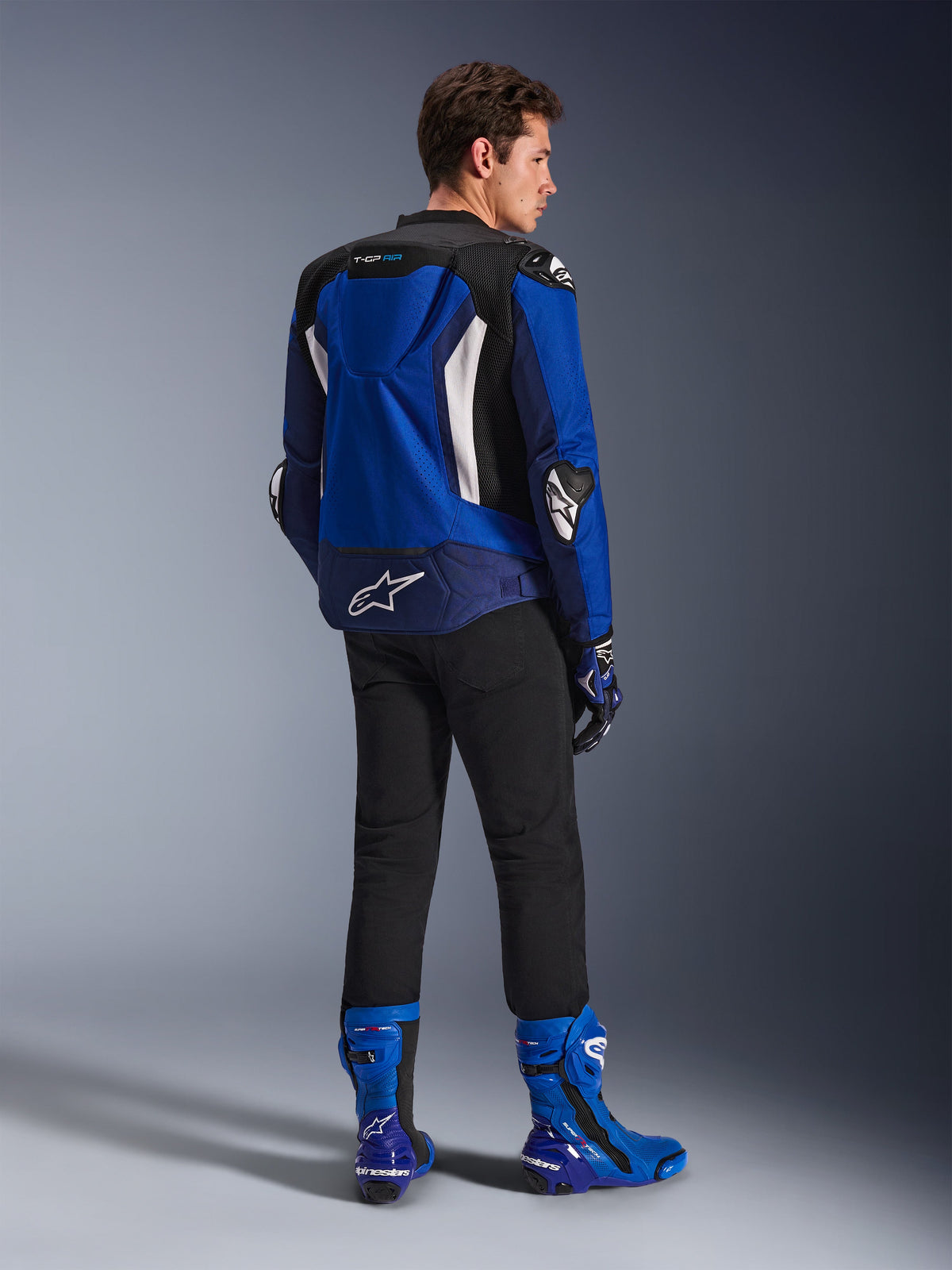 Alpinestars T-GP Air-jacka, sporttextiljacka, Bright Blue Navy White, sedd bakifrån på en modell som bär svarta byxor och matchande blå och vita stövlar, med vit Alpinestars-logotyp på nedre ryggen, ventilationspaneler i mesh och en aerodynamisk puckel
