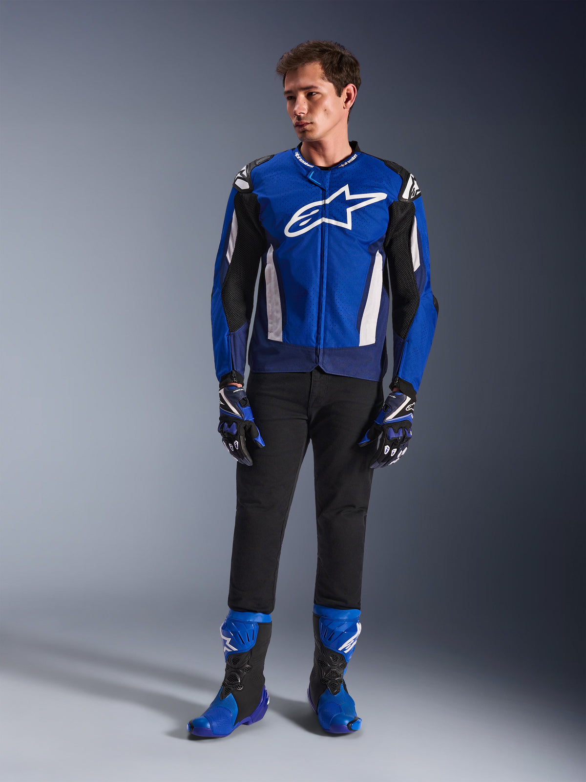 Manlig modell som bär Alpinestars T-GP Air-jacka i Bright Blue Navy White, matchad med svarta byxor, matchande blå och marinblå racinghandskar och blå och svarta motorcykelstövlar, studiobild som visar en komplett outfit för sportkörning.