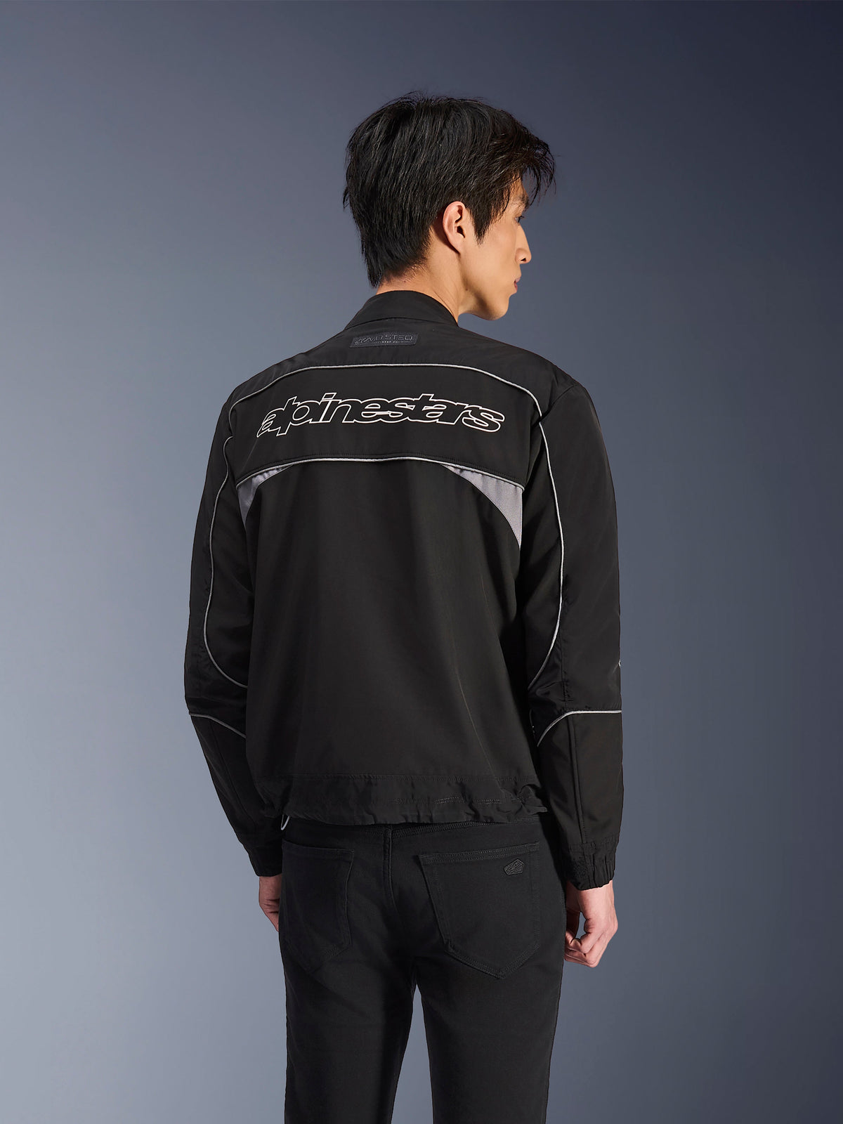 Alpinestars Unite-jacka, urban motorcykeljacka, svart med mörkgrå accenter, vy bakifrån som visar vit Alpinestars-logotyp över axlarna, reflekterande passpoaler och paneler, minimalistisk softshell-design i streetwear-stil