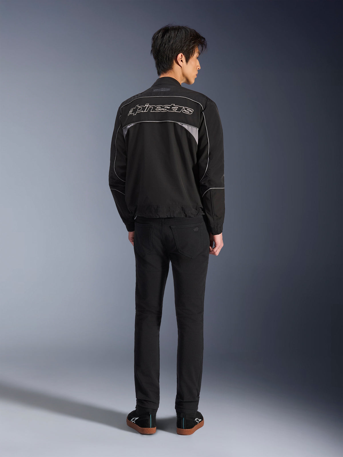 Alpinestars Unite-jacka, svart och mörkgrå motorcykeljacka, vy bakifrån som visar Alpinestars konturlogotyp på ryggen, grå reflekterande paneler, minimalistisk urban design, bärs med svarta byxor och svarta skor.