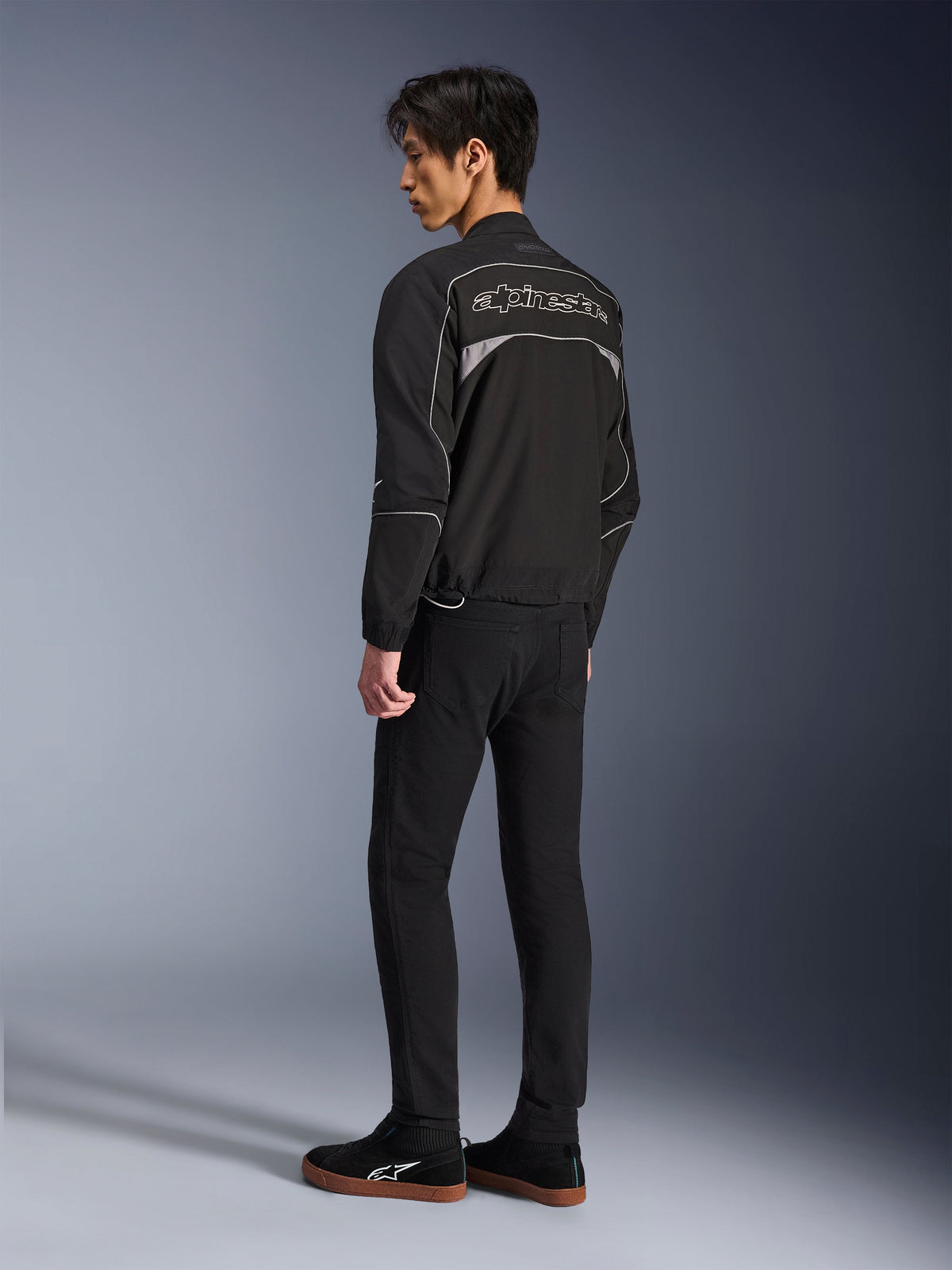 Alpinestars Unite-jacka, urban motorcykeljacka, svart och mörkgrå, baksida som visar en stor konturerad Alpinestars-logotyp, reflekterande piping och paneler, minimalistisk softshell-design, bärs med svarta byxor och vardagliga skor