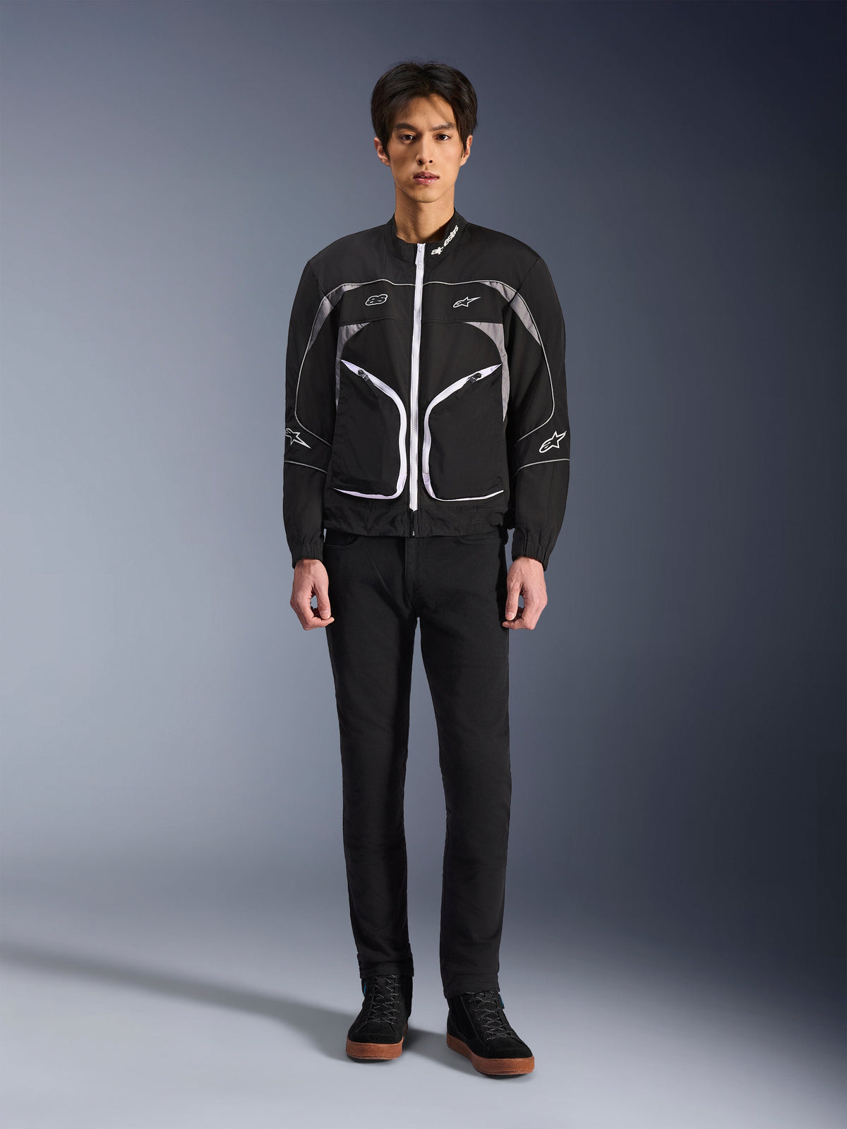 Alpinestars Unite-jacka, motorcykeljacka, svart och mörkgrå, bärs av en modell med svarta byxor och svarta stövlar, med vita kontrasterande dragkedjor, reflekterande detaljer och minimalistisk urban design