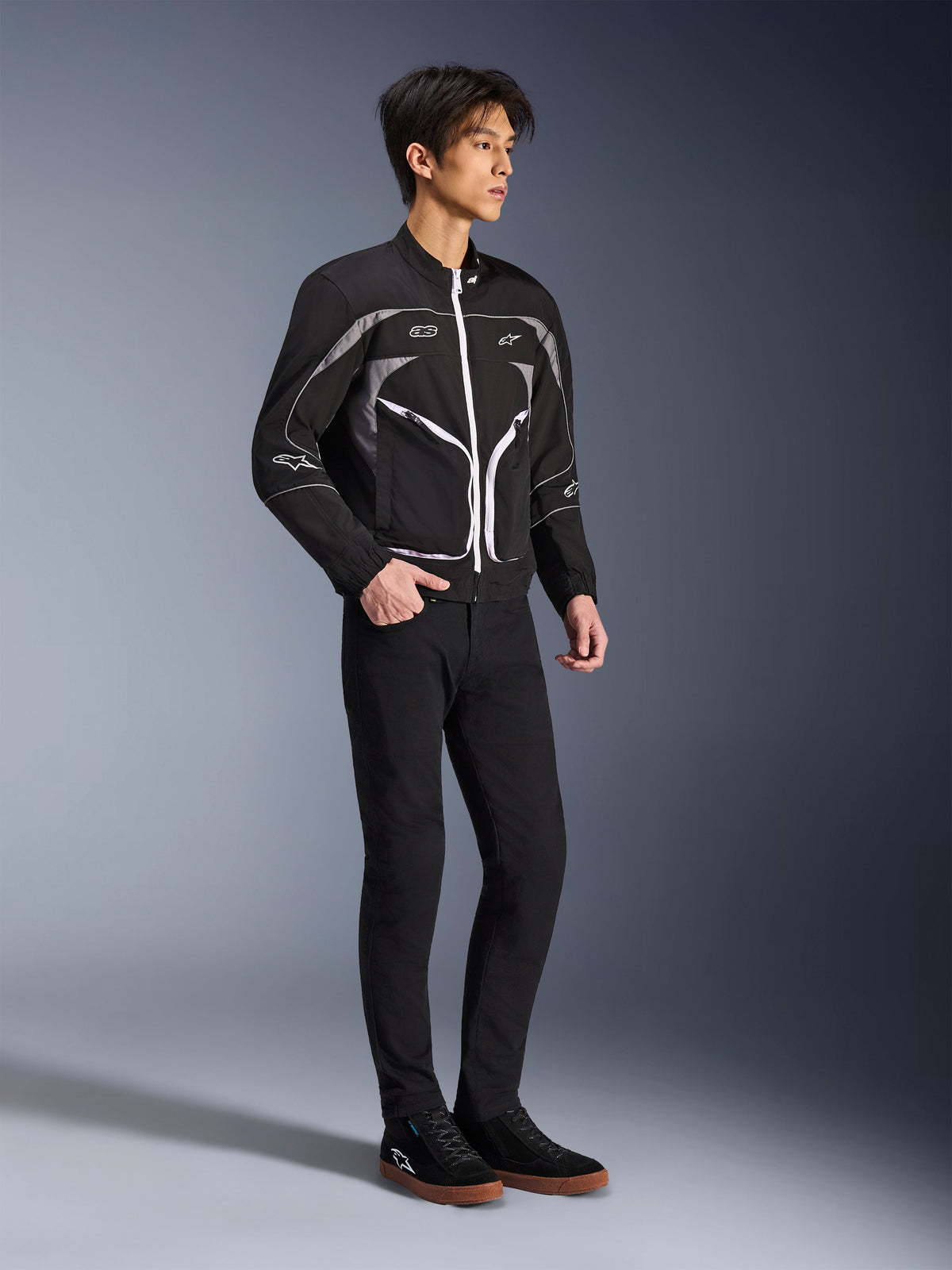 Modell som bär Alpinestars Unite Jacket i svart och mörkgrått, urban motorcykeljacka med vita dragkedjor och reflekterande detaljer, kombinerad med svarta slim-fit-byxor och sneakers, minimalistisk gatuinspirerad körutrustning.