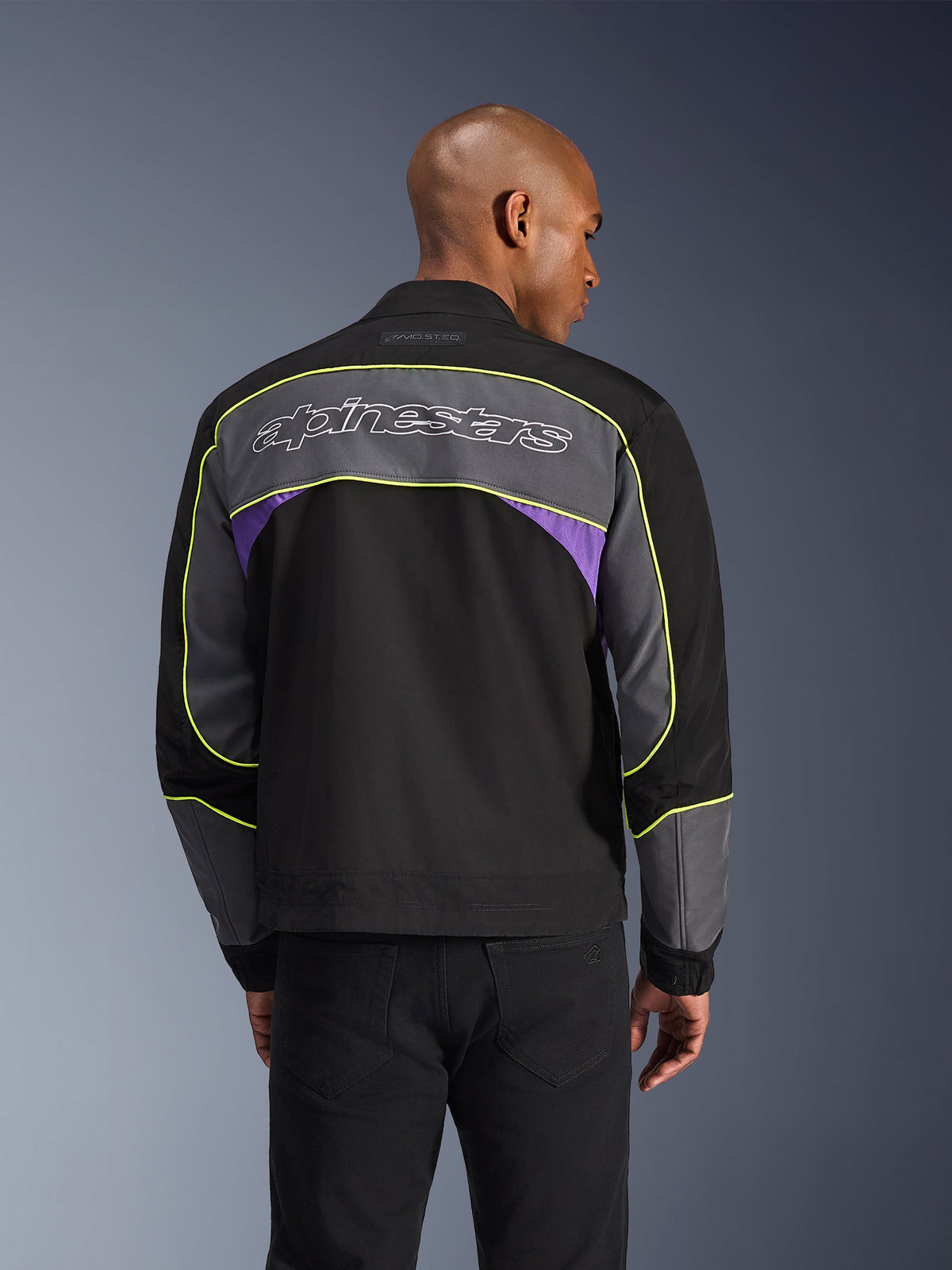 Alpinestars Unite-jacka, urban motorcykeljacka, Black Asphalt Violet, ryggvy med grå paneler och violetta accenter, limegul piping, stor Alpinestars-text på övre ryggen, minimalistisk streetwear-design