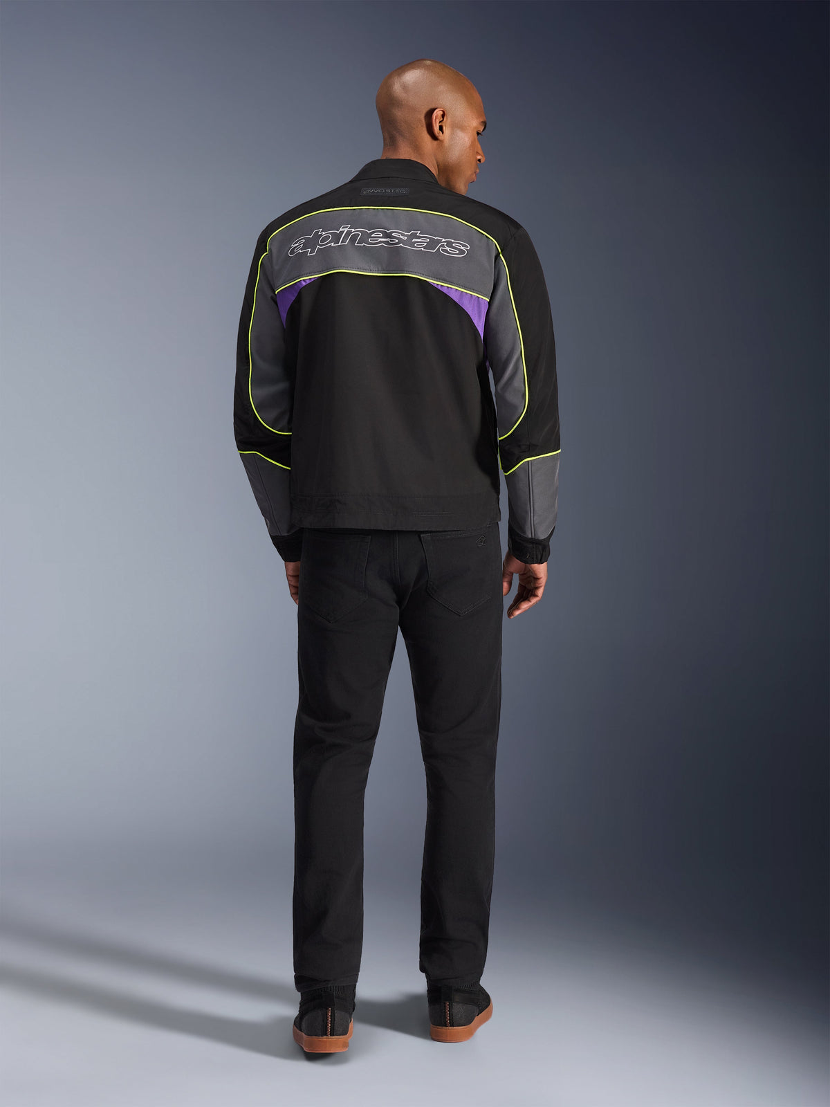 Alpinestars Unite-jacka, urban motorcykeljacka i Black Asphalt Violet, vy bakifrån som visar grå ryggpaneler med lila accenter och neongula passpoaler, Alpinestars-logotyp i skrivstil över axlarna, bärs med svarta byxor.