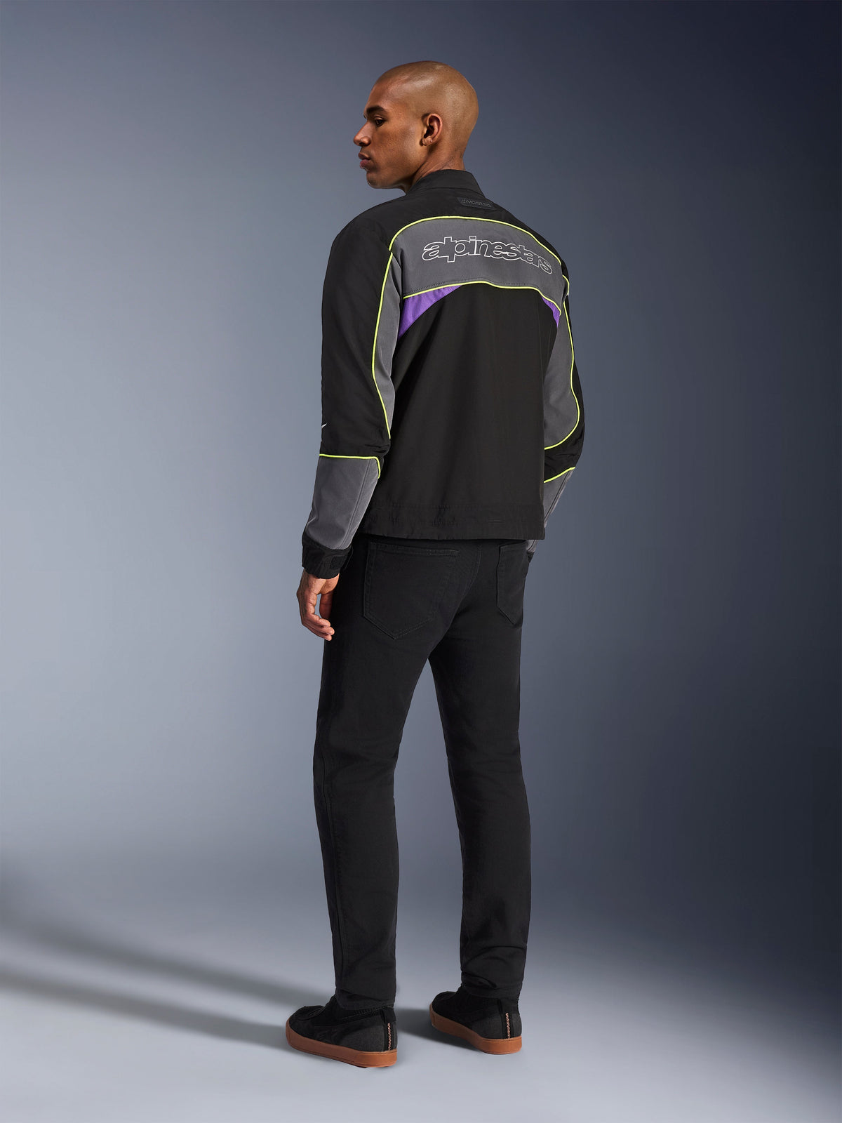Alpinestars Unite-jacka, urban motorcykeljacka, Black Asphalt Violet, trekvartsvy bakifrån på modell, svart softshell med grå paneler och lila accenter, neongula reflekterande detaljer, Alpinestars-logotyp på övre delen av ryggen, matchad med svarta byxor och svarta vardagsskor