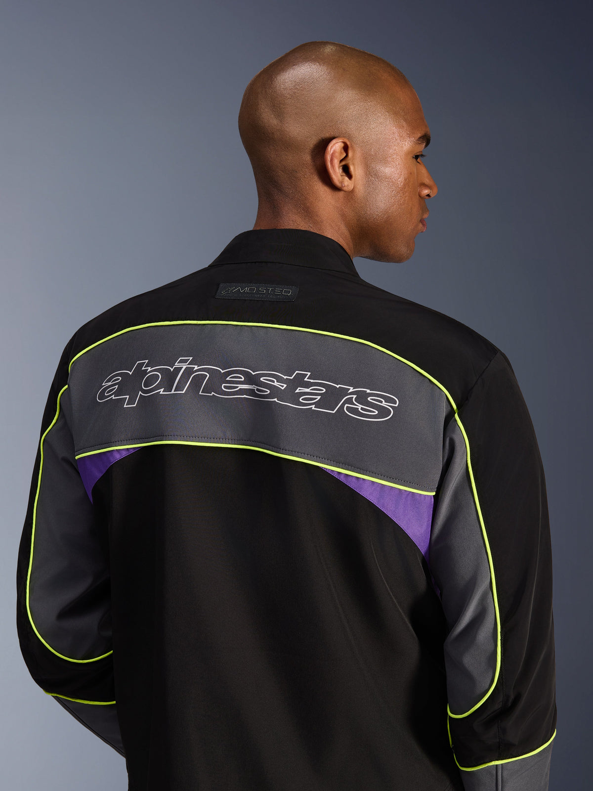 Alpinestars Unite-jacka, urban motorcykeljacka, Black Asphalt Violet, ryggvy som visar en stor grå axelpanel med Alpinestars-logotyp, neongula kantband och violetta accenter på en 3-lagers softshell-konstruktion