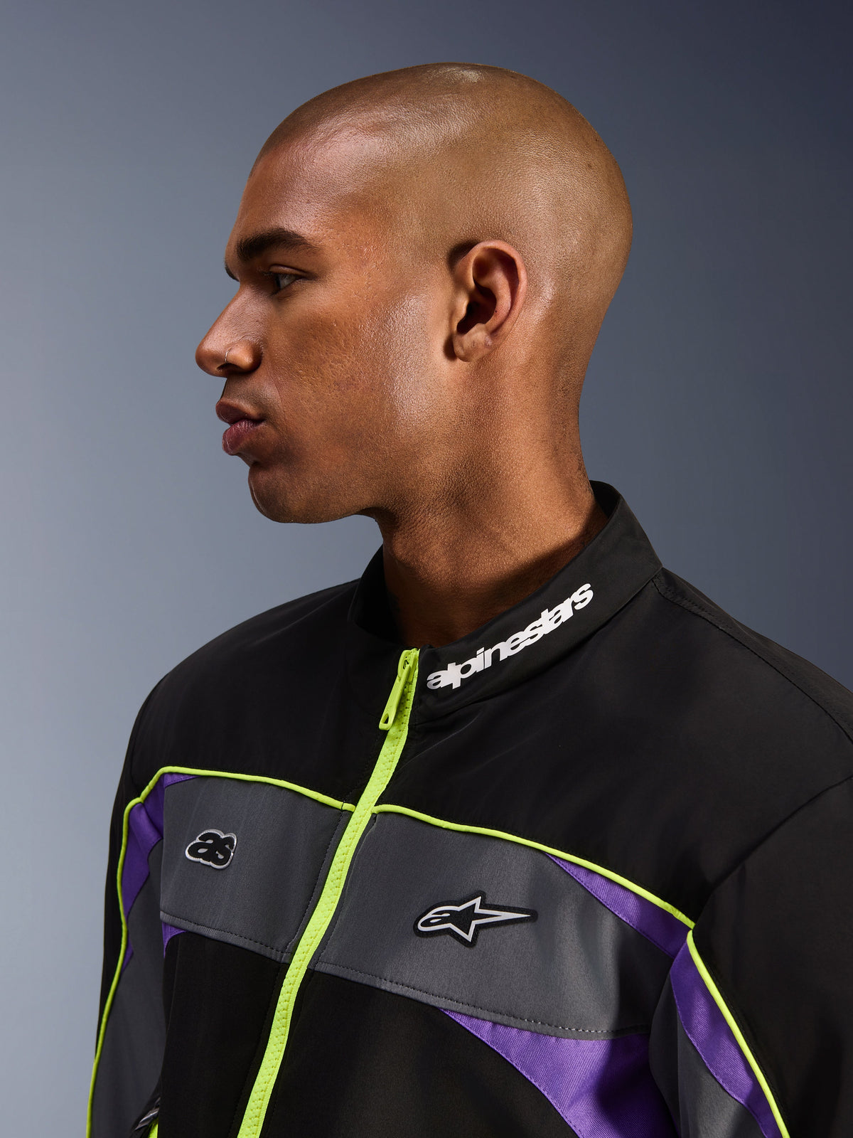 Alpinestars Unite-jacka, Black Asphalt Violet, profilvy av manlig modell med fokus på den höga kragen med vit Alpinestars-logotyp, neongul dragkedja, grå bröstpaneler med logotyper, lila detaljer, minimalistisk urban streetwear-stil för motorcykel