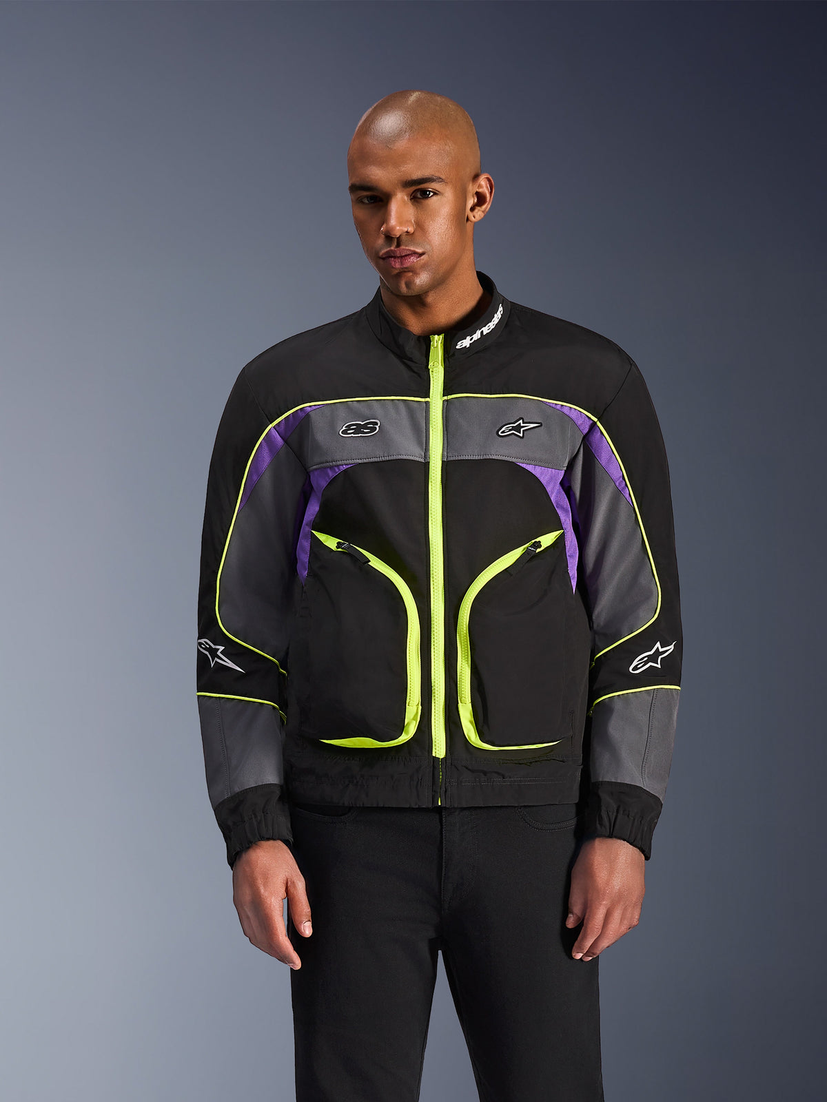 Alpinestars Unite Jacket, urban motorcykeljacka, Black Asphalt och lila med neongula detaljer, stora framfickor med neongula dragkedjor, hög krage med vit logotyp, bärs av modell, modern streetwear-inspirerad skyddsutrustning