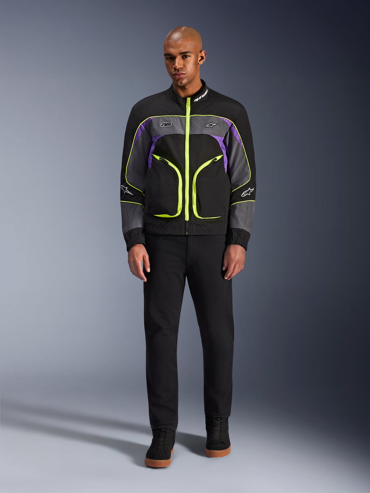 Manlig modell som bär Alpinestars Unite Jacket i Black Asphalt Violet, med en svart bas, grå och lila paneler och livfulla neongula dragkedjor och detaljer, kombinerat med svarta byxor och skor, minimalistisk urban motorcykeljacka.