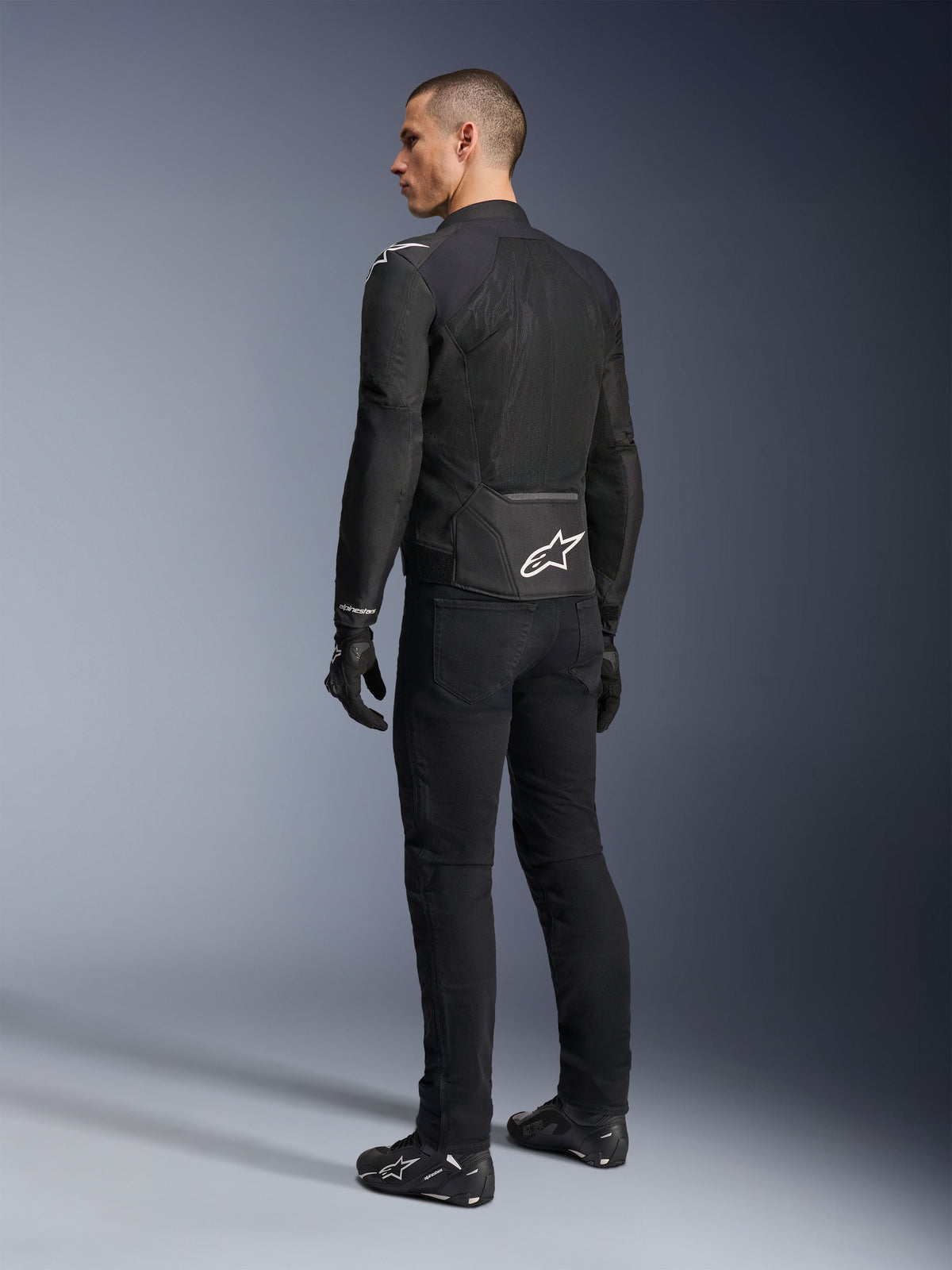 Alpinestars Viper Air V4-jacka, svart motorcykeljacka i textil, bakifrånvy på en manlig modell, med stora paneler i teknisk mesh för luftflöde och en vit stjärnlogotyp på nedre delen av ryggen, buren med matchande svarta körbyxor och handskar
