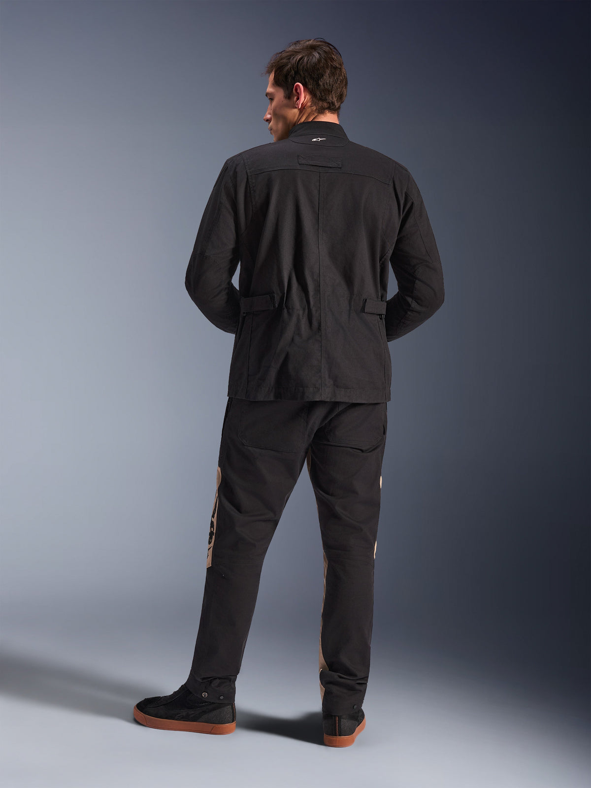 Alpinestars Dice Jacket i svart sedd bakifrån, urban motorcykeljacka i fältmodell med veckade axlar och midjejusteringar, matchad med svarta och sandfärgade mc-byxor och sneakers, modern stadspendlarstil