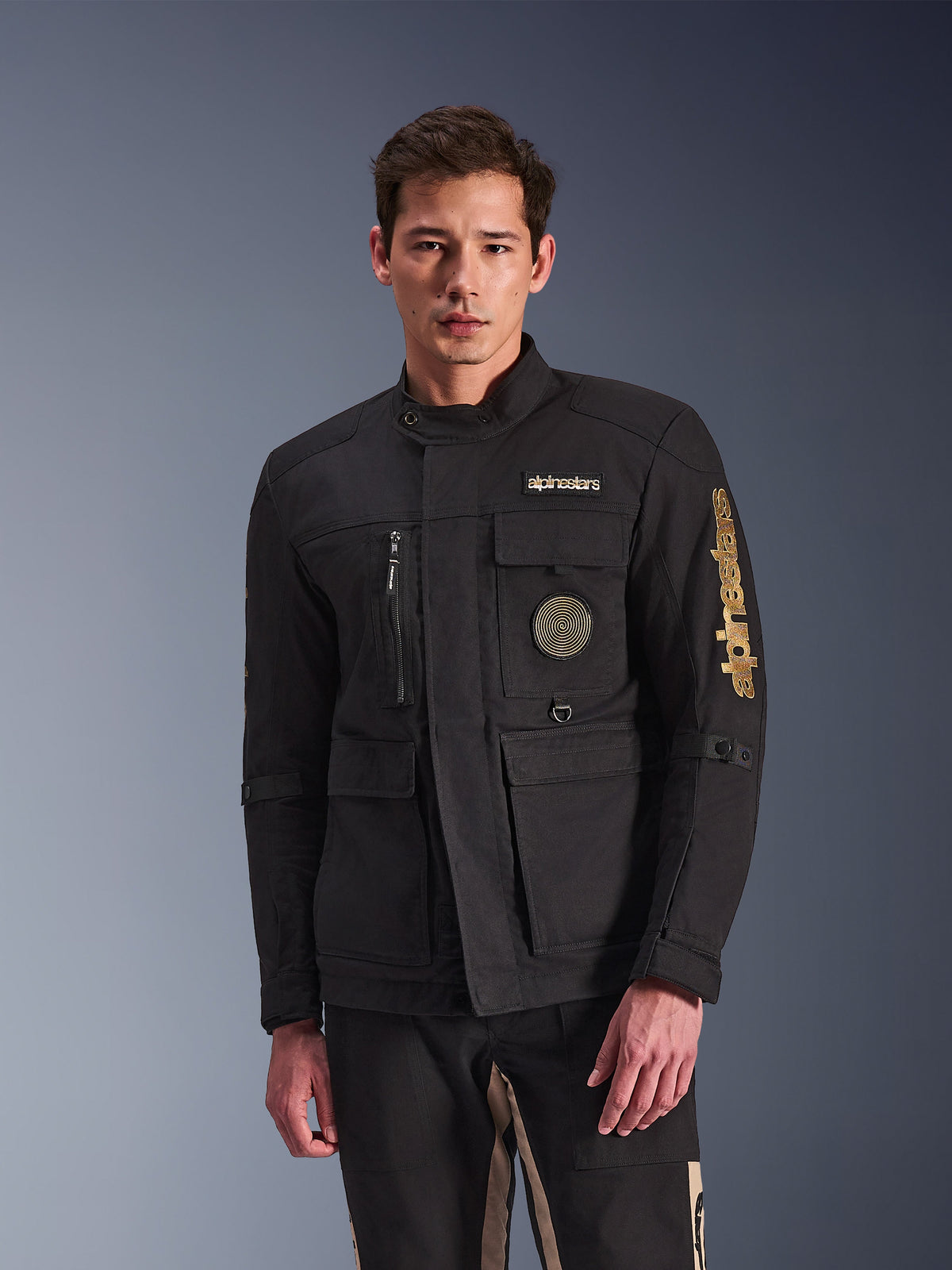 Alpinestars Dice Jacket, svart motorcykeljacka, bärs av modell, med guldlogotyper från Alpinestars, flera funktionella fickor, cirkulärt spiralmärke och design i urban fältjackestil