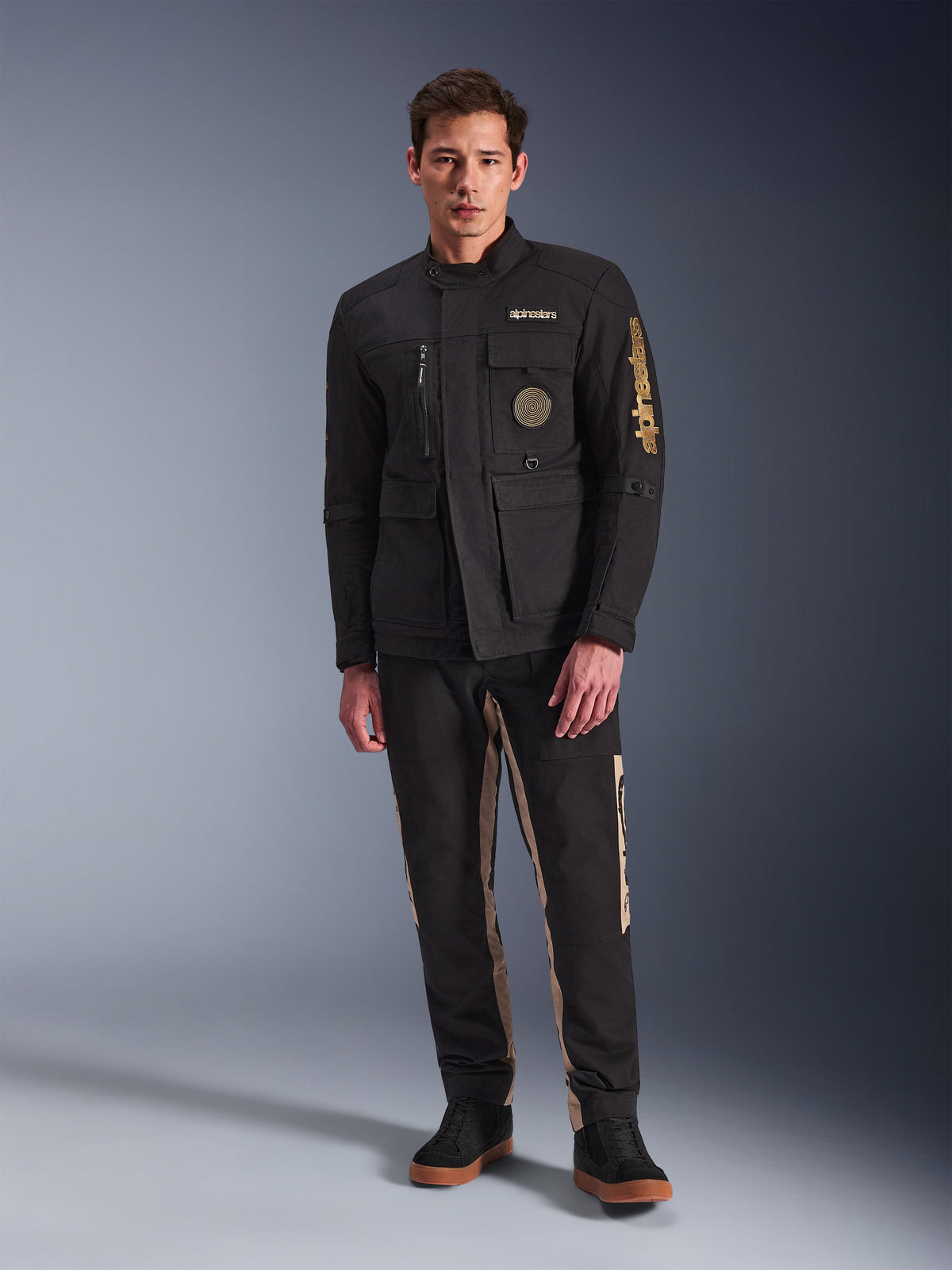 Manlig modell i Alpinestars Dice-jacka, svart urban motorcykeljacka med vintage fältjacka-stil, fyra funktionella framfickor, Alpinestars-logotyp på ärmen och bröstet, kombinerad med svarta och beige byxor, modern urban körutrustning