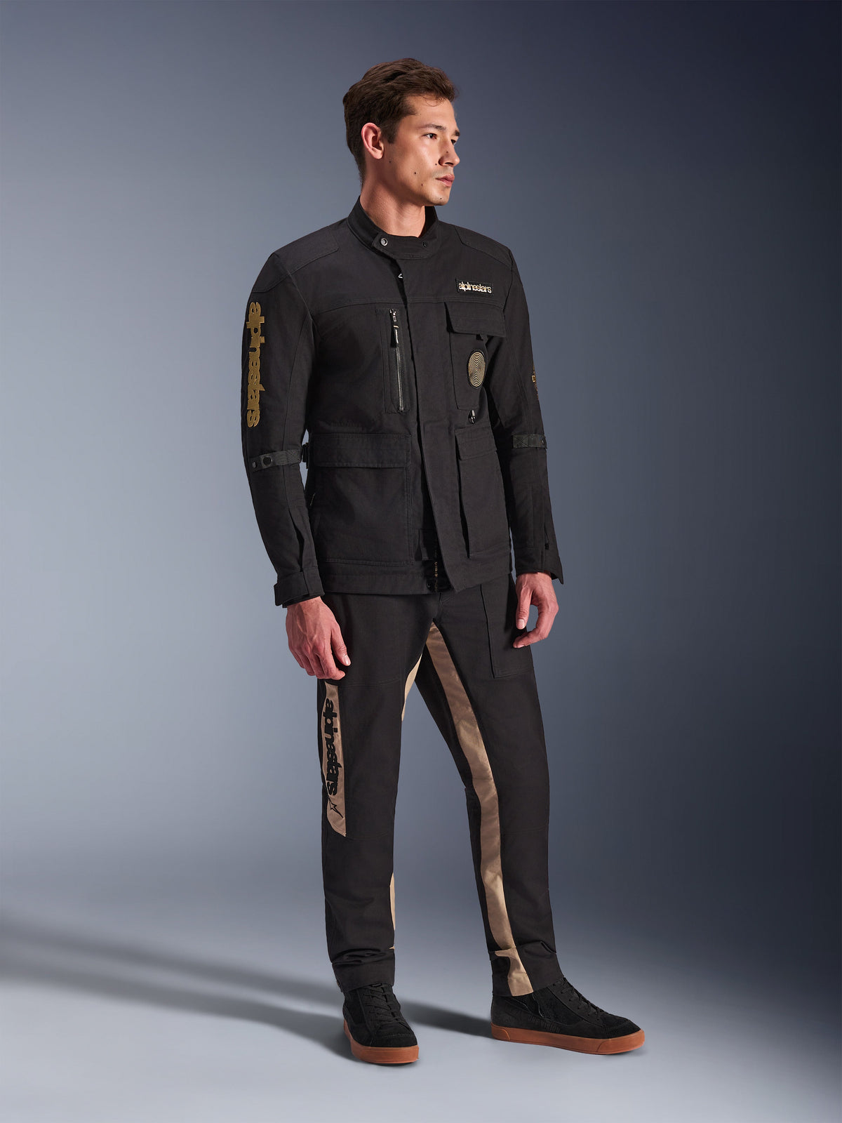 Manlig modell bär Alpinestars Dice-jacka i svart, urban motorcykeljacka med vintage fältjacka-estetik, utrustad med flera praktiska fickor och guldlogotyp på ärmen, kombinerat med matchande svarta och beigea byxor och svarta sneakers.