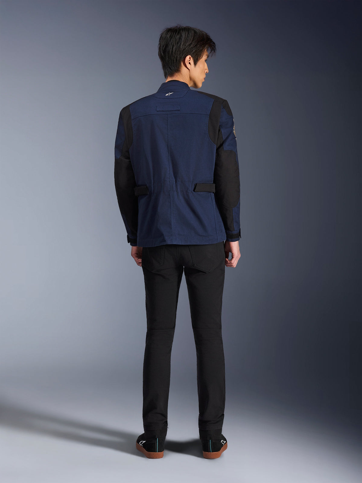 Modell som bär Alpinestars Dice-jackan i Dark Blue och Black, vy bakifrån, urban mc-fältjacka med boxveckad axelkonstruktion och justerbara flikar i midjan, matchat med svarta byxor och svarta sneakers.