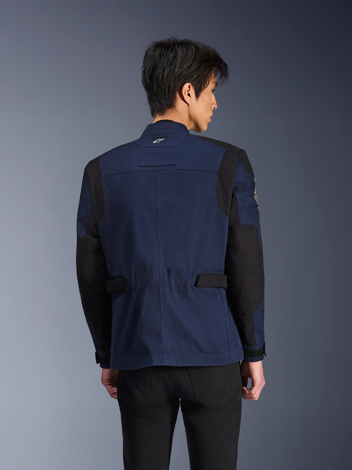Alpinestars Dice Jacket, mörkblå och svart urban fältjacka för motorcykel, sedd bakifrån bärs av en man, med förstärkta svarta axel- och armbågspaneler och justerbara flikar i midjan.