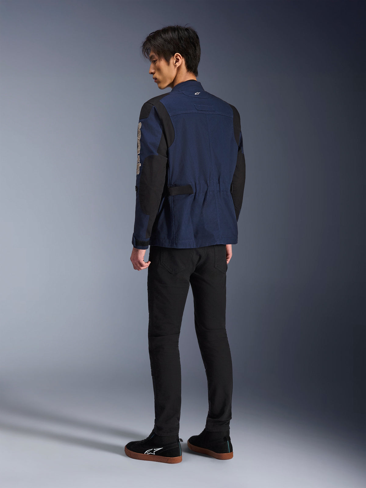 Manlig modell sedd bakifrån iklädd Alpinestars Dice Jacket i mörkblått och svart, kombinerad med svarta byxor och svarta sneakers, urban motorcykel-fältjacka med boxveckade axlar och justerbara midjeflikar.