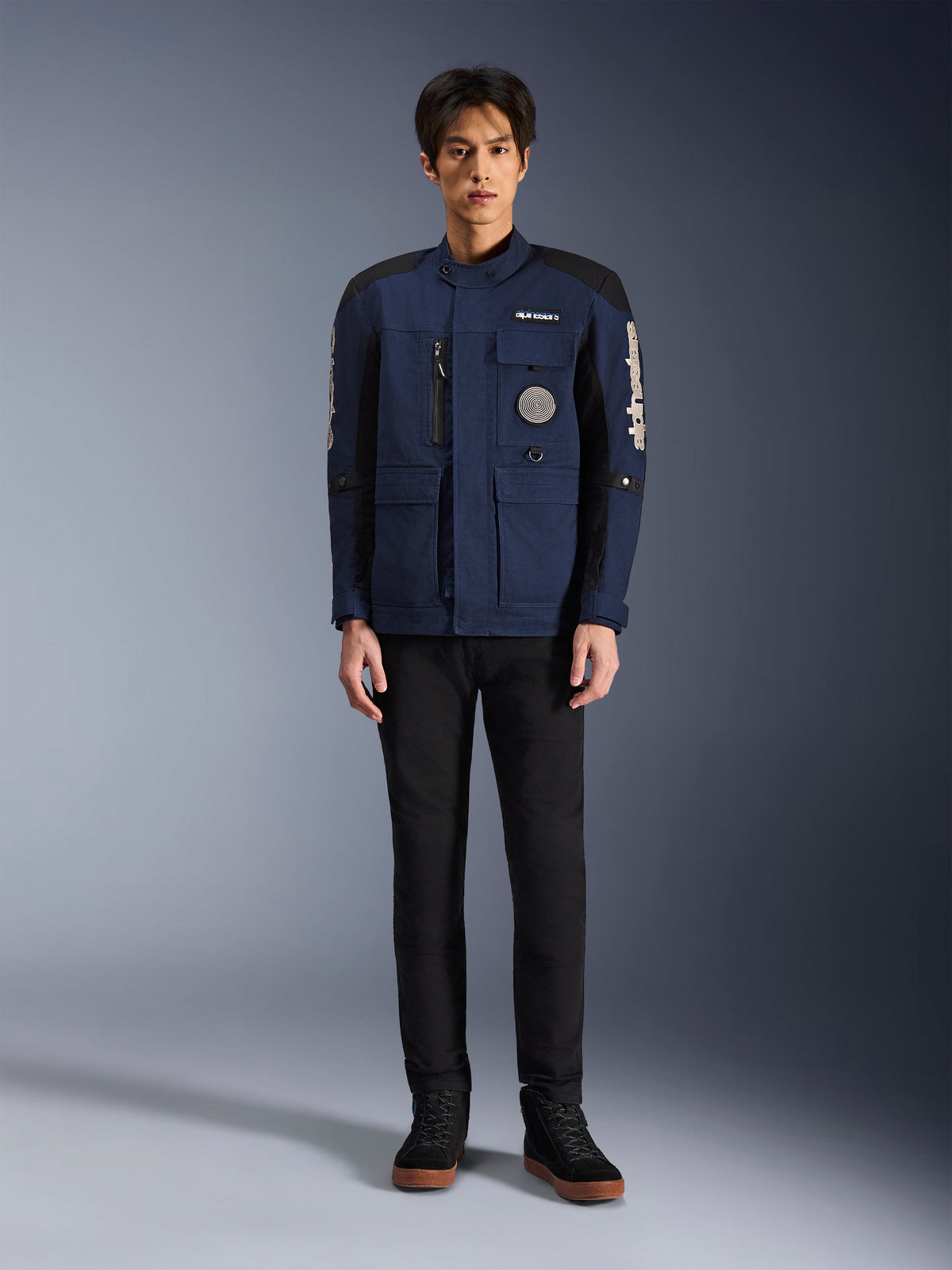 Alpinestars Dice-jacka, urban motorcykeljacka, mörkblå och svart, bärs av en modell med svarta byxor och svarta skor, modern fältjackdesign med flera fickor, vertikal dragkedja på bröstet och vit Alpinestars-logotyp på ärmarna.