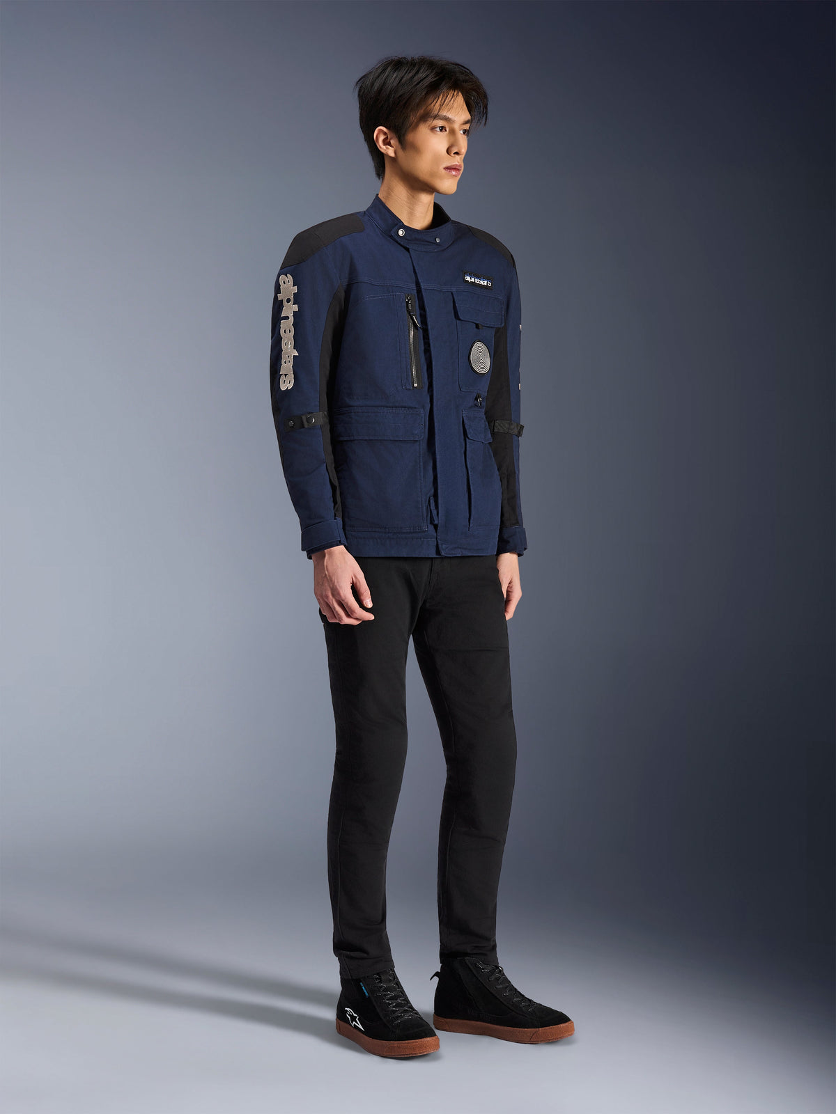 Alpinestars Dice Jacket, mörkblå och svart, modern motorcykeljacka i fältmodell som bärs av en modell med svarta byxor och svarta high-top-sneakers, med flera funktionella fickor, vertikal Alpinestars-logotyp på ärmen och vävda märken, urban modern körutrustning
