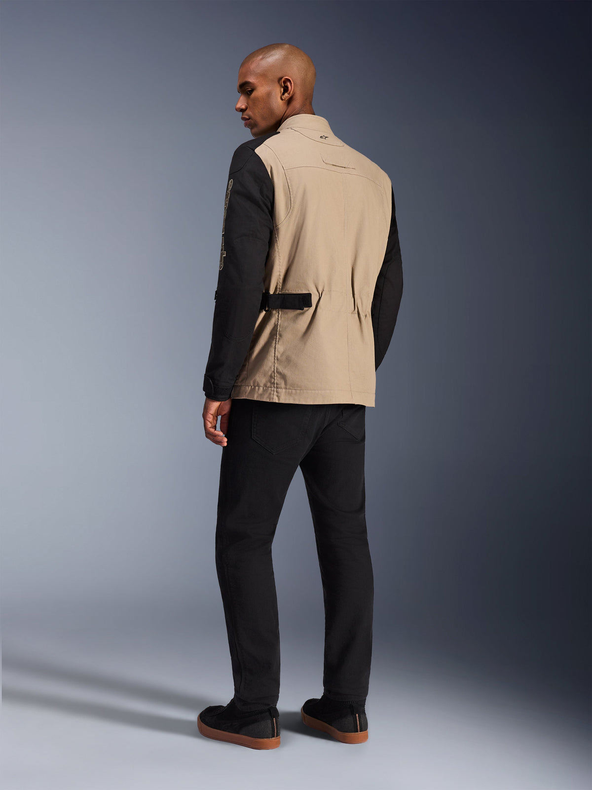 Alpinestars Dice-jacka, urban motorcykeljacka, Aluminum Black med beige bål och svarta ärmar, sedd bakifrån på modell, robust arbetsklädesinspirerad design med midjejusteringar, matchad med svarta körbyxor.